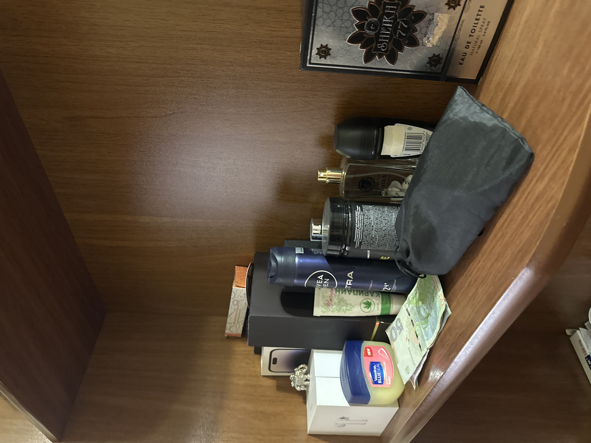 A cluttered shelf contains various items. From top to bottom: A box of "Sheikh 77" eau de toilette, a black toiletry bag, several bottles of perfume, a can of "Nivea Men" deodorant, a tube labeled "КАРИПАИН" and "VEA EN TRA," a box appearing to contain a phone (possibly an iPhone), a small decorative object, a container of Vaseline, a box containing what appear to be Apple AirPods, and some Polish 50 złoty banknotes. The shelf itself appears to be made of wood.