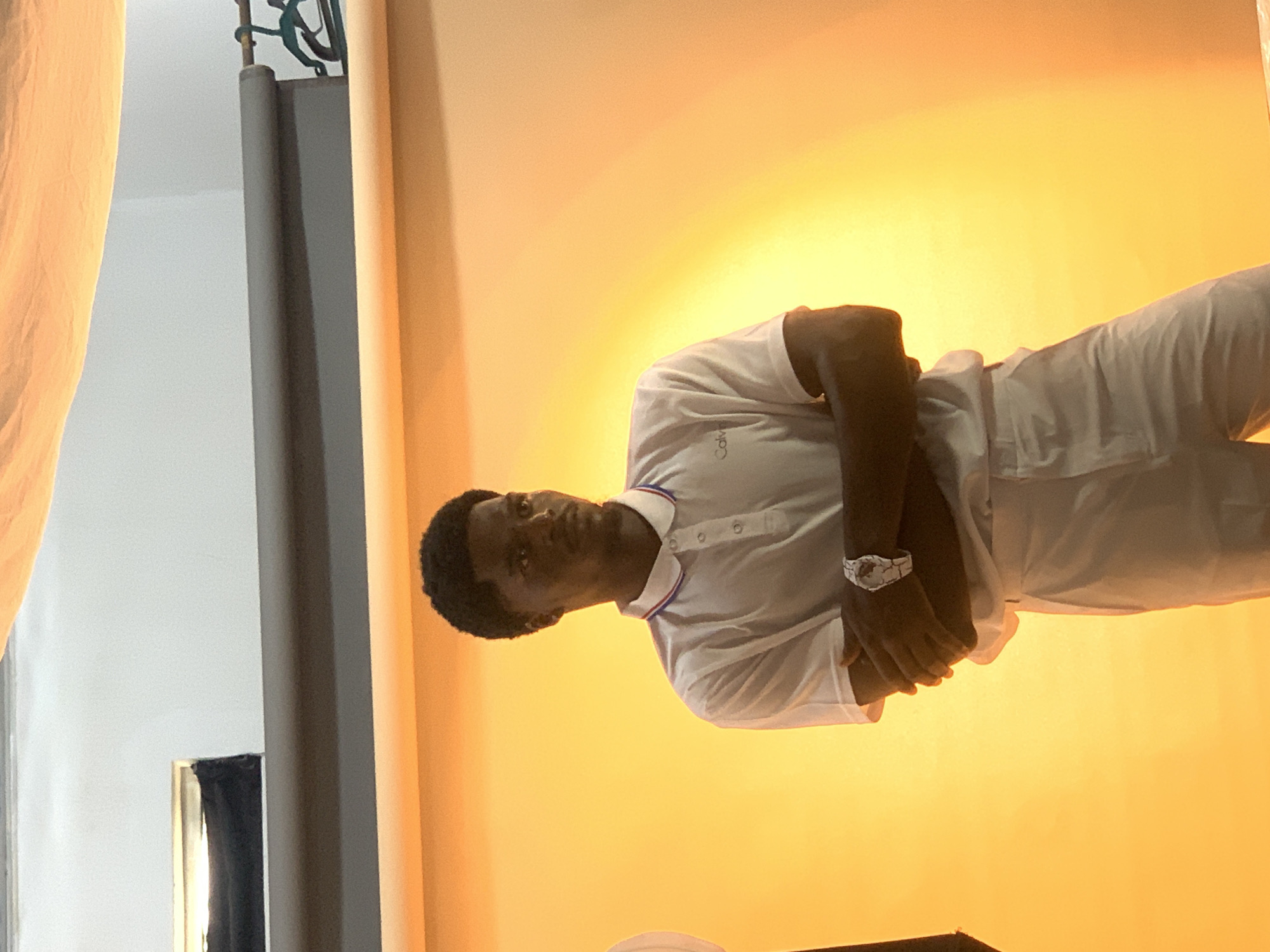 The image is a portrait of a dark-skinned man with short hair, wearing a white short-sleeved polo shirt and white pants. He's standing in front of a backdrop that transitions from a light orange to a bright yellow. He has a watch on his left wrist. The left side of the image features a grey metal frame and a partially visible white curtain. The location is Gura Top, Nigeria.