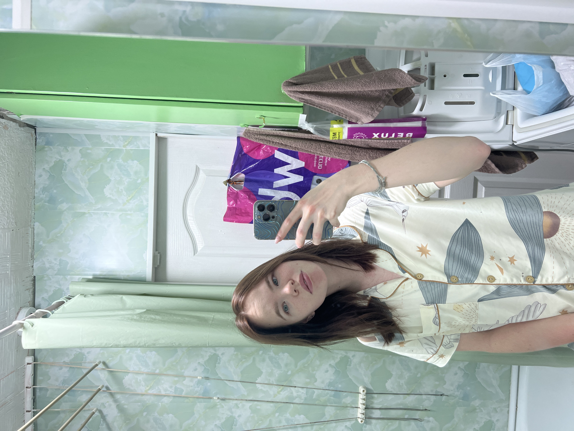A young woman with shoulder-length brown hair and blue eyes is taking a mirror selfie in a bathroom, her expression neutral. She is wearing cream-colored pajamas adorned with a pattern of abstract gray leaf-like shapes, golden stars, and crescent moons, finished with brown piping and buttons. Her left hand holds a dark smartphone, its multiple camera lenses visible, while a braided bracelet is on her wrist.
The mirrored background reveals a modest bathroom in Chaikovsky, Russia. Above the woman, a green shelf or cabinet is reflected. To her left, a white door with decorative panels stands next to a wall covered in light green tiles or wallpaper featuring a swirling, marble-like pattern. Below her, a white clothes drying rack with horizontal bars is mounted on the wall, and a light green shower curtain or similar fabric hangs from a rod above it. To her upper right, inside a reflected cabinet or shelf, several small brown towels are seen alongside a purple package displaying the reversed text "XULƎB," which reads "BEJUX." Other plastic bags are also stored within the cabinet. The scene captures an ordinary moment, likely in the morning or evening given the pajamas.