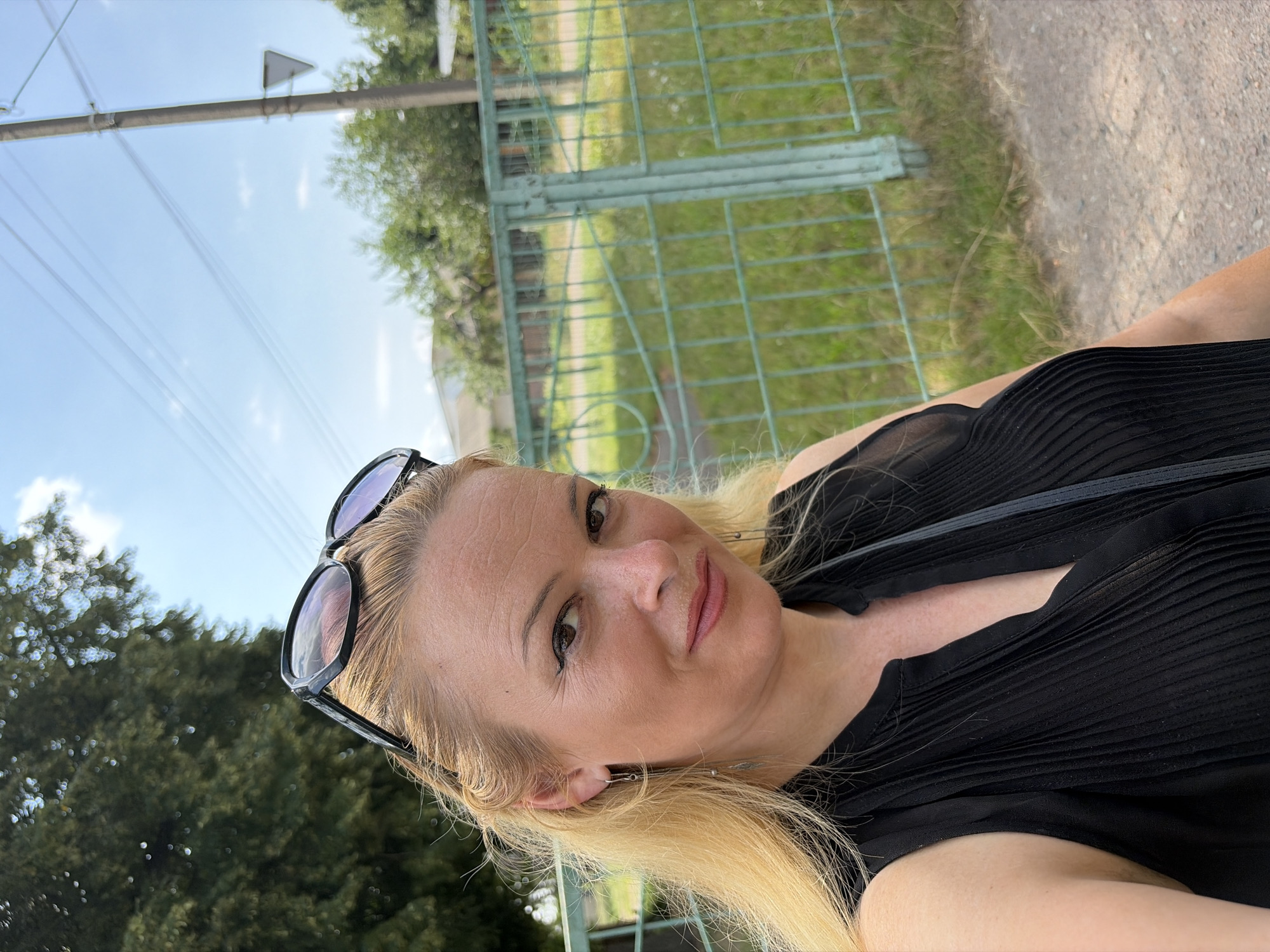 A woman with blonde hair and sunglasses on her head takes a selfie outdoors. She is wearing a black sleeveless top with pleats. She has on dangling earrings. Behind her is a green fence and trees. The sky is blue with some clouds.