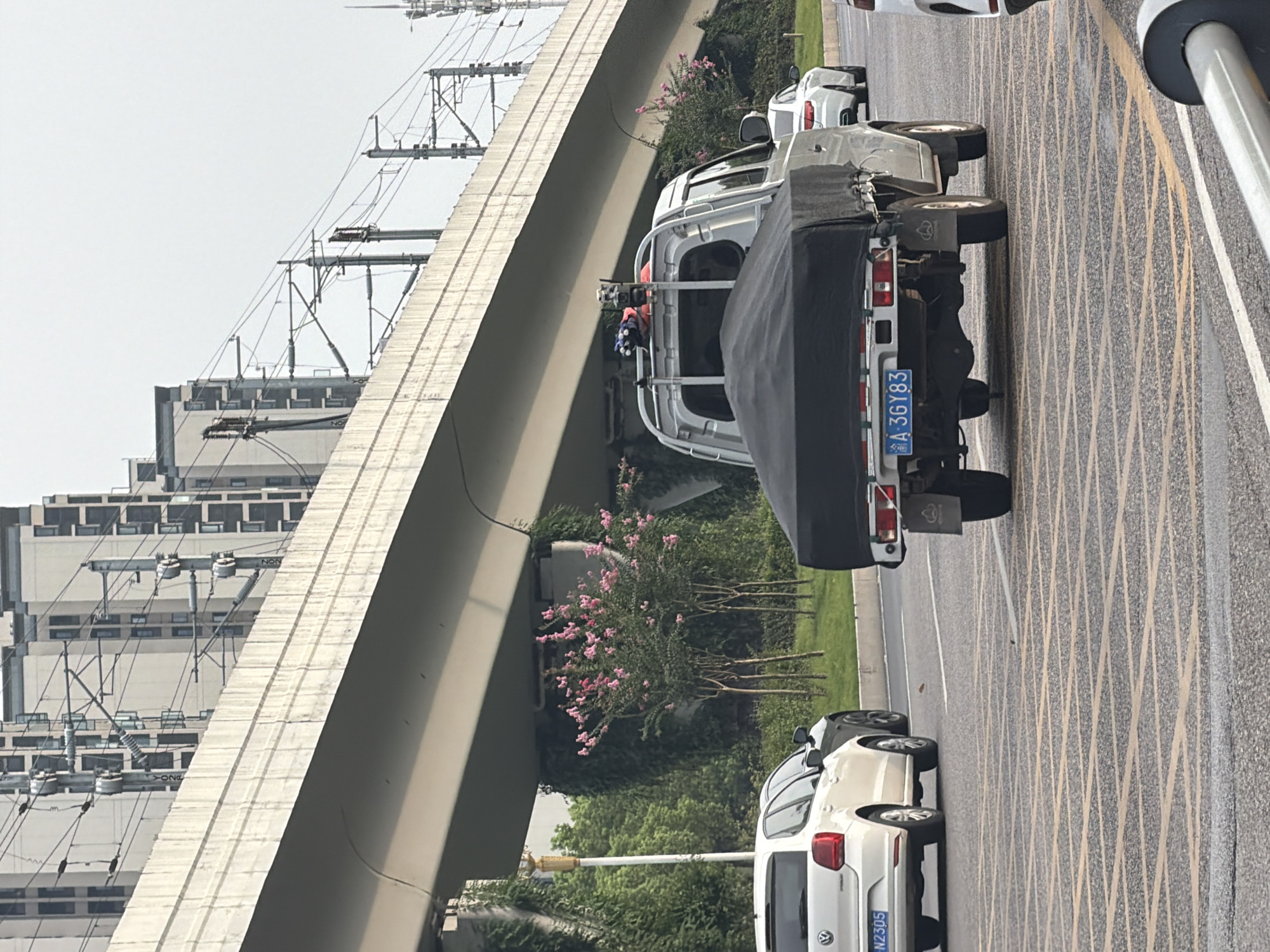 In Jiangjin, China, a silver pickup truck with a black tarp covering its bed is driving on a street under a large elevated structure. The truck has a blue license plate that reads "A 3GY83". A white Volkswagen Polo is to the left of the truck, and a white sedan is to the right. The street is marked with yellow grid lines. Greenery and pink flowering trees are visible along the side of the road, and tall buildings and overhead power lines are in the background.
