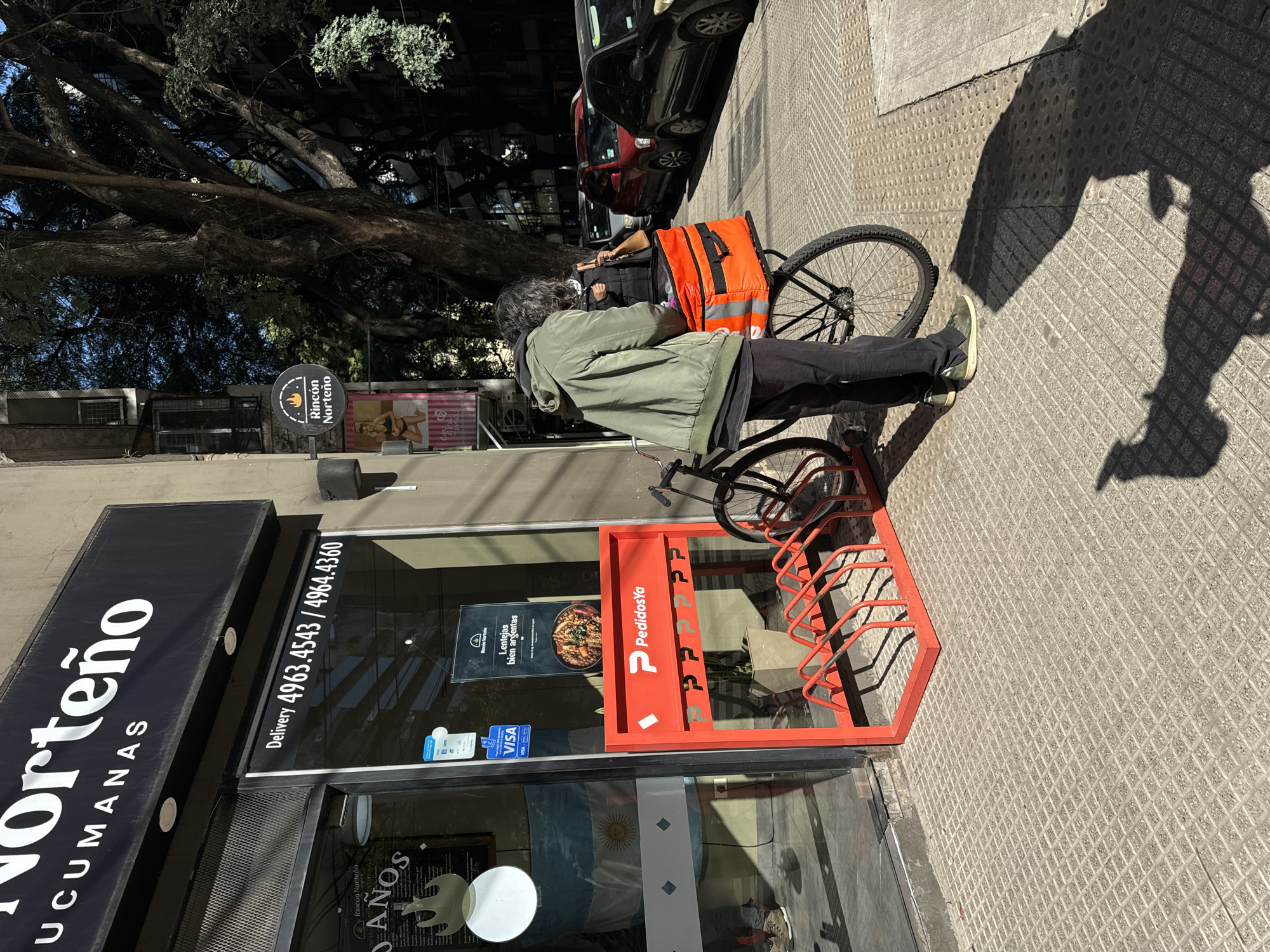 A medium shot captures a delivery cyclist standing next to their bicycle, parked in front of a restaurant. The cyclist, dressed in an olive green jacket and dark pants, wears an orange PedidosYa delivery backpack. The bicycle, equipped with a rear rack, is parked beside a bright red PedidosYa bicycle rack positioned against the restaurant's facade. The restaurant, named "Norteño Tucumanas," has a black sign with white lettering and a delivery phone number displayed on the window: "Delivery 4963.4543 / 4964.4360." A smaller sign reads "Rincon Norteño" above "10 ANOS" (10 Years). Inside the restaurant, a flag of Argentina is visible behind a glass window. A menu board featuring "Lentejas bien argentas" (Argentinian Lentils) is also displayed on the window. The image suggests the location is in Buenos Aires, Argentina, based on the presence of the Argentinian flag. The street has textured concrete tiles, and a shadow cast by a tree or building suggests it is daytime. A car can be seen parked nearby in the background.