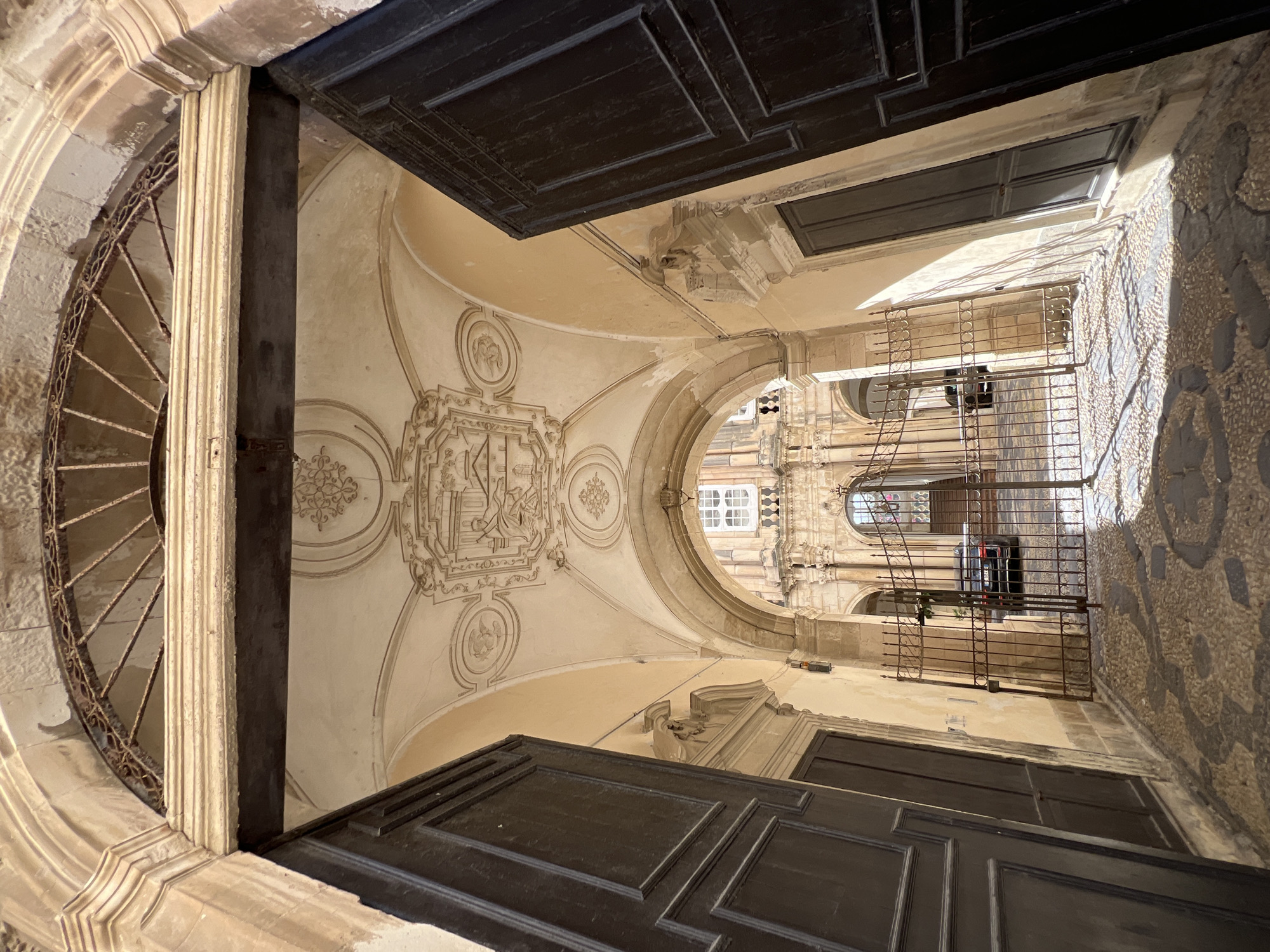 An arched entranceway, lined with dark wooden doors on either side, leads into a sun-drenched courtyard. The ceiling of the entrance features ornate plasterwork and a coat of arms. Beyond the entrance, a wrought-iron gate is visible, beyond which a dark-colored car is parked in a courtyard paved with cobblestones. Sunlight streams into the courtyard, casting long shadows. The architecture suggests a historic location, likely within the city of Syracuse, Italy, possibly in an area with Baroque influences.