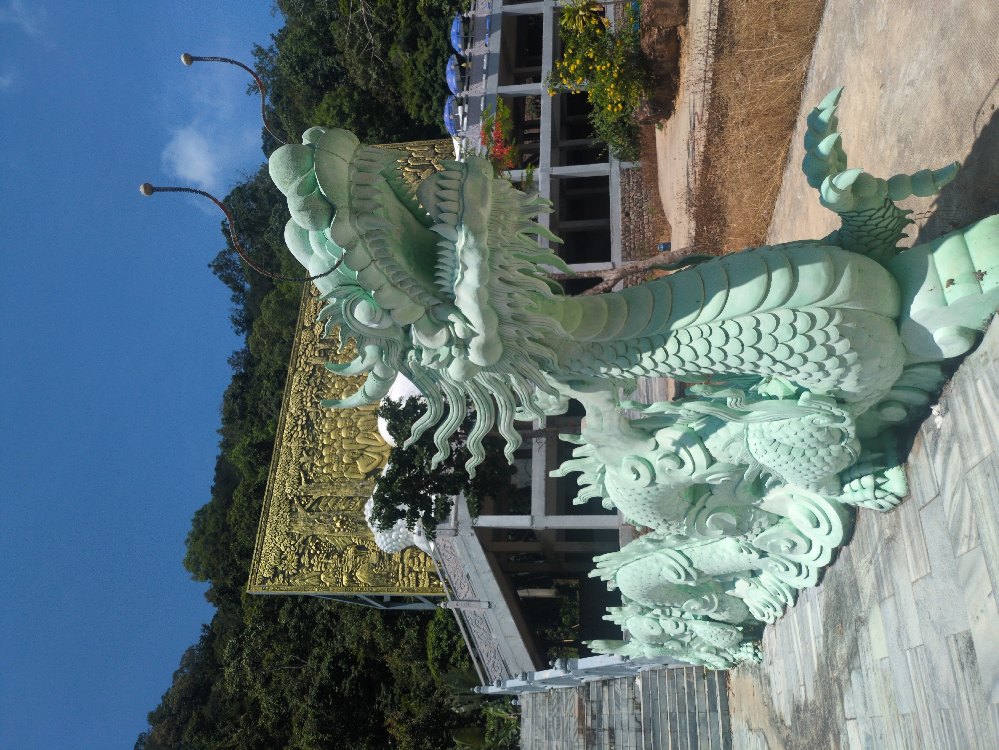 A low-angle, outdoor, daytime shot shows a light-green, large dragon statue with bug-like antenna atop a stairway and stone walkway, with a gold colored wall structure and trees in the background, in Phú Quốc, Vietnam.
The dragon statue is positioned on the right side of the image and angled from the viewer toward the left side of the image. The dragon has an open mouth with fangs, a long mane, scaly body, and clawed feet. Below the dragon statue's body, additional aquatic statues are positioned. The walkway underneath the dragon statue has stairs with metal railings on the left side of the image.
Behind the left side of the dragon statue's head, a gold colored wall structure covered in people and nature artwork is visible. Trees fill the background behind the wall structure. On the upper-right side of the image, a white building, trees, and dry vegetation are visible.
The sky is light blue and clear on the left side of the image. The shot is in portrait format, with the top of the image oriented toward the left side of the view.