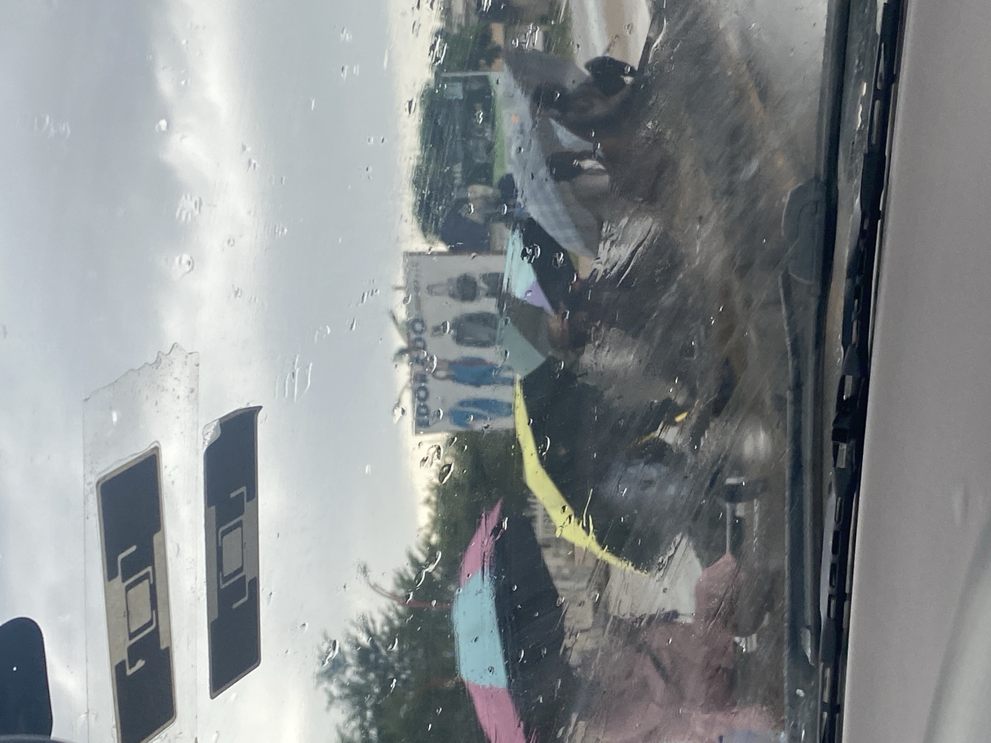 The image captures a scene through a rain-streaked windshield, suggesting the perspective of someone inside a vehicle. In the foreground, the windshield wipers are visible at the bottom. Through the raindrops, a group of people holding umbrellas can be seen walking. The umbrellas are of various colors, including pink, blue, and yellow. In the background, a billboard displays images of people, possibly models, wearing clothing items like jackets. Trees and a cloudy sky are also visible, indicating an outdoor environment. The overall impression is of a rainy day in a city. While the image context suggests Keffi, Nigeria, this is not directly evident from visual cues within the image itself.