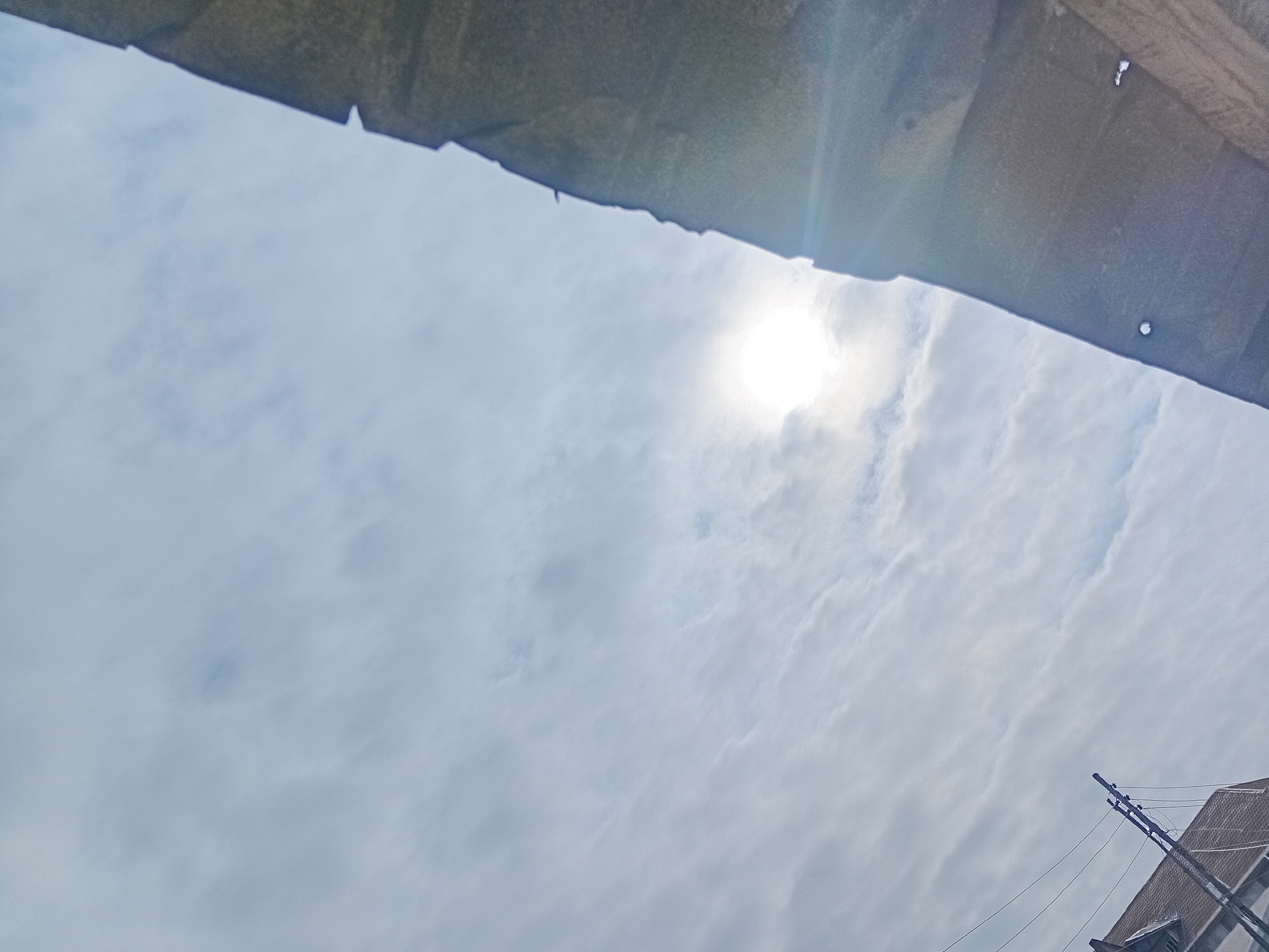 An upward angle shot shows a cloudy sky with the sun visible as a bright orb. In the lower right corner, the top of a building with a red roof is visible, along with electrical poles and wires. A dark, textured overhang extends across the top of the frame, casting a shadow that cuts across the image diagonally.