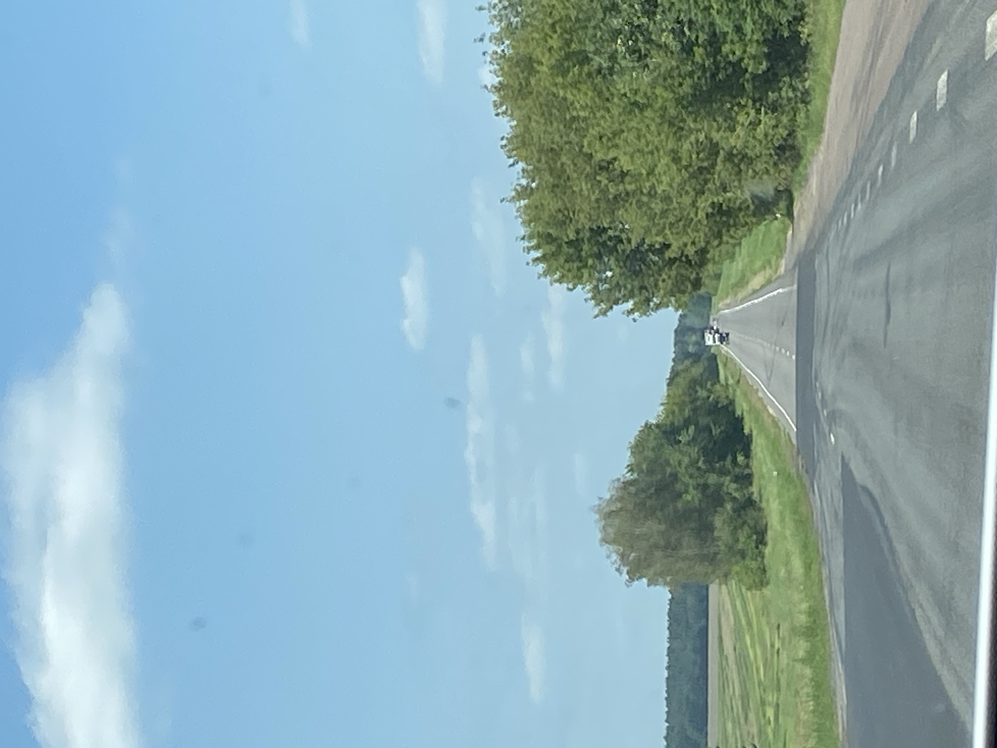 The image depicts a view from a vehicle traveling on a paved road through a rural landscape. The sky is bright blue with scattered white clouds. On the left side of the road, there is a vast green field with mown grass arranged in rows, bordered by a dense line of trees in the distance. On the right side of the road, a row of lush green trees lines the route. Several vehicles, including what appears to be a white truck and a smaller white vehicle, are visible further down the road. The road itself is asphalt with white lane markings, and there are some dark patches on the surface, possibly due to heat or moisture. The overall scene suggests a bright, sunny day in a rural area of Belarus, likely within the Матыкальскі сельскі Савет given the provided context.