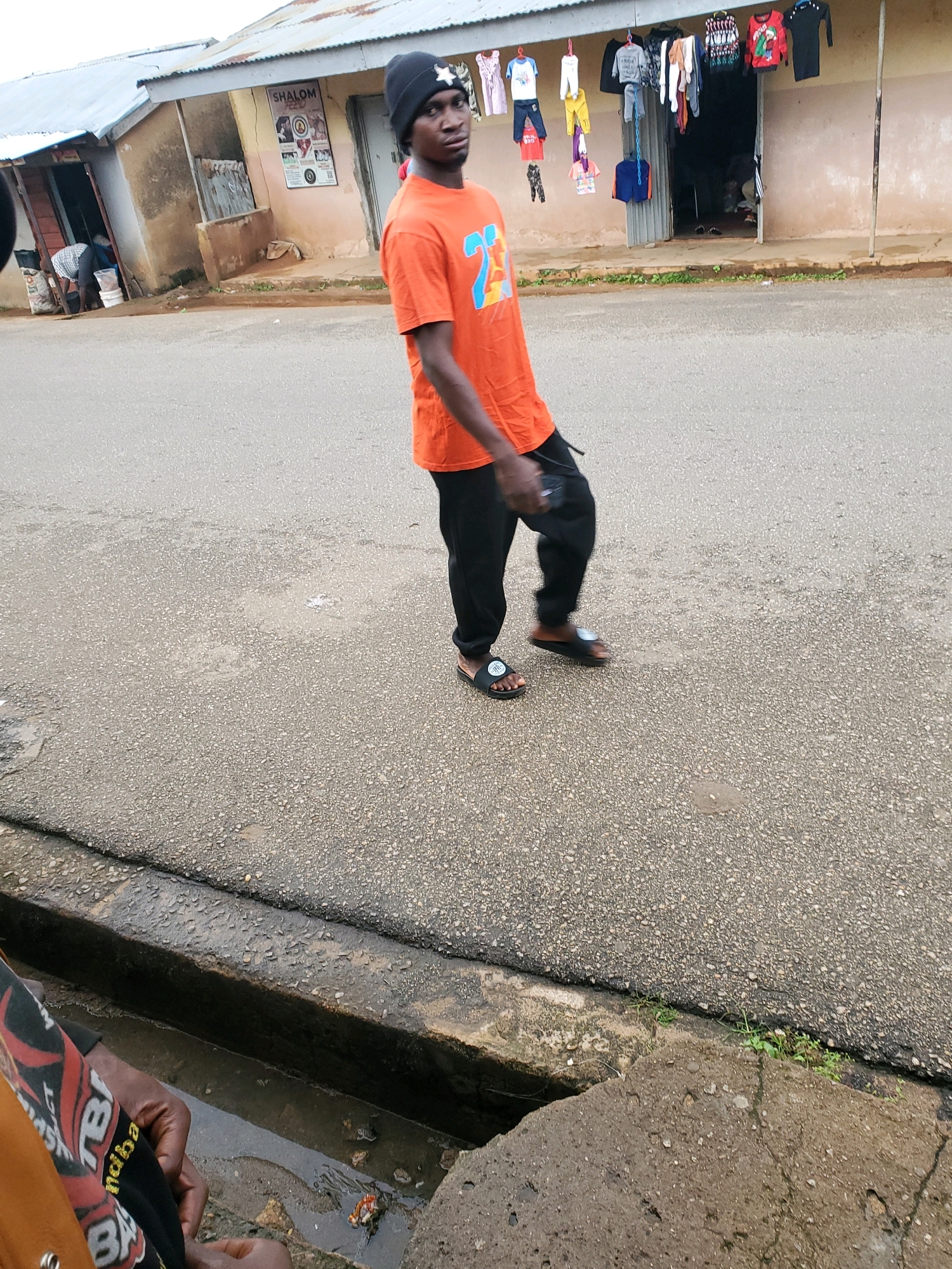 A young Black man wearing an orange t-shirt with the number "27" on it and black pants walks on a paved street in front of a building with shops. He has a black beanie on his head with a white star on it. The building has a metal roof and clothes are hanging on a line in front of one of the shops. A drainage ditch runs along the side of the street.