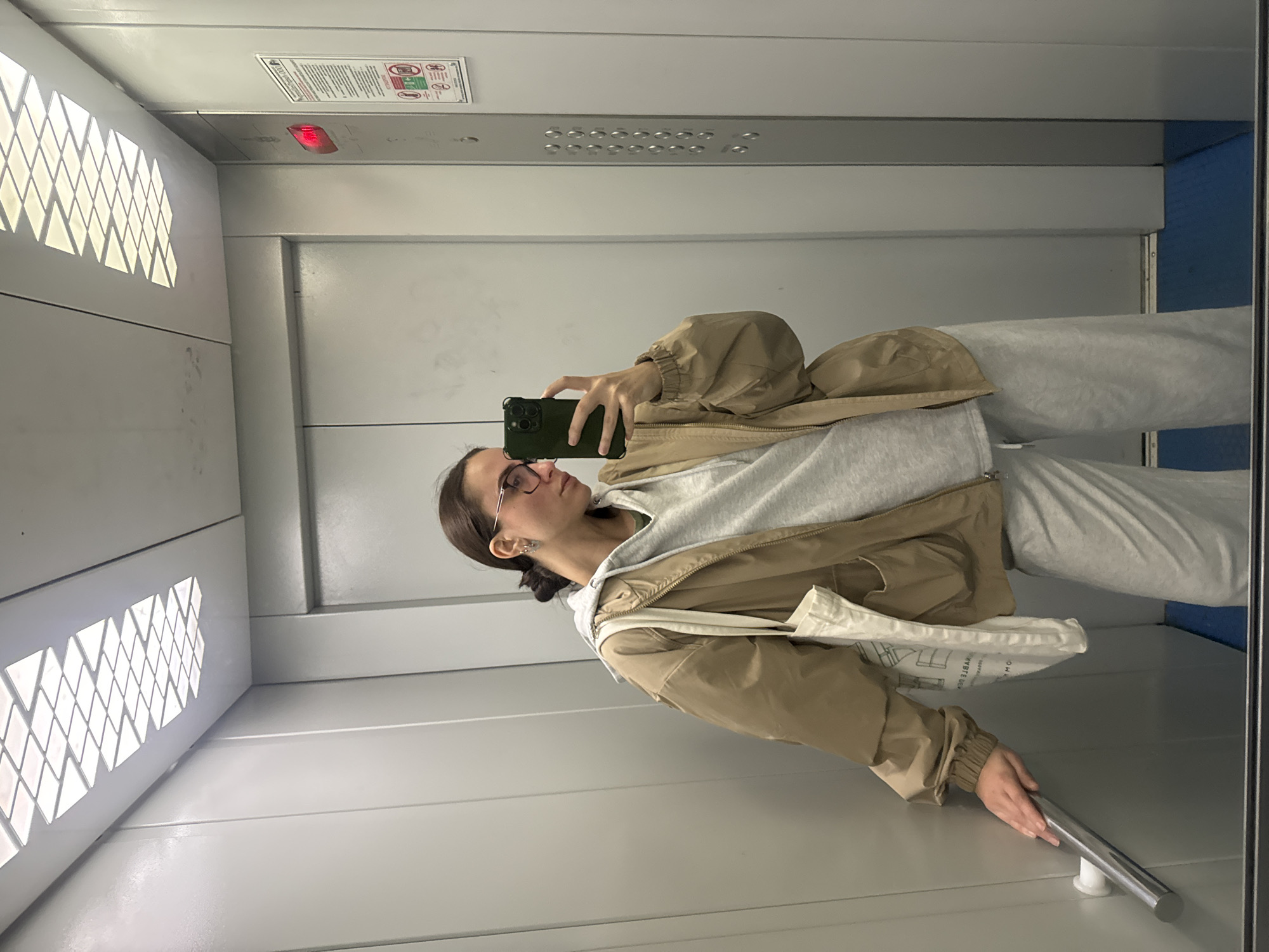 A woman is taking a selfie in an elevator. She is wearing glasses, a grey hoodie, and a beige jacket, and she is holding a smartphone in her left hand. She is standing in front of a mirror, reflecting her image and the elevator interior. The elevator has a panel with buttons and a light indicating the current floor. The walls are grey, and the lighting is bright. A sign with instructions is affixed to the upper wall. The location is likely in a building in Krasnoyarsk, Russia.