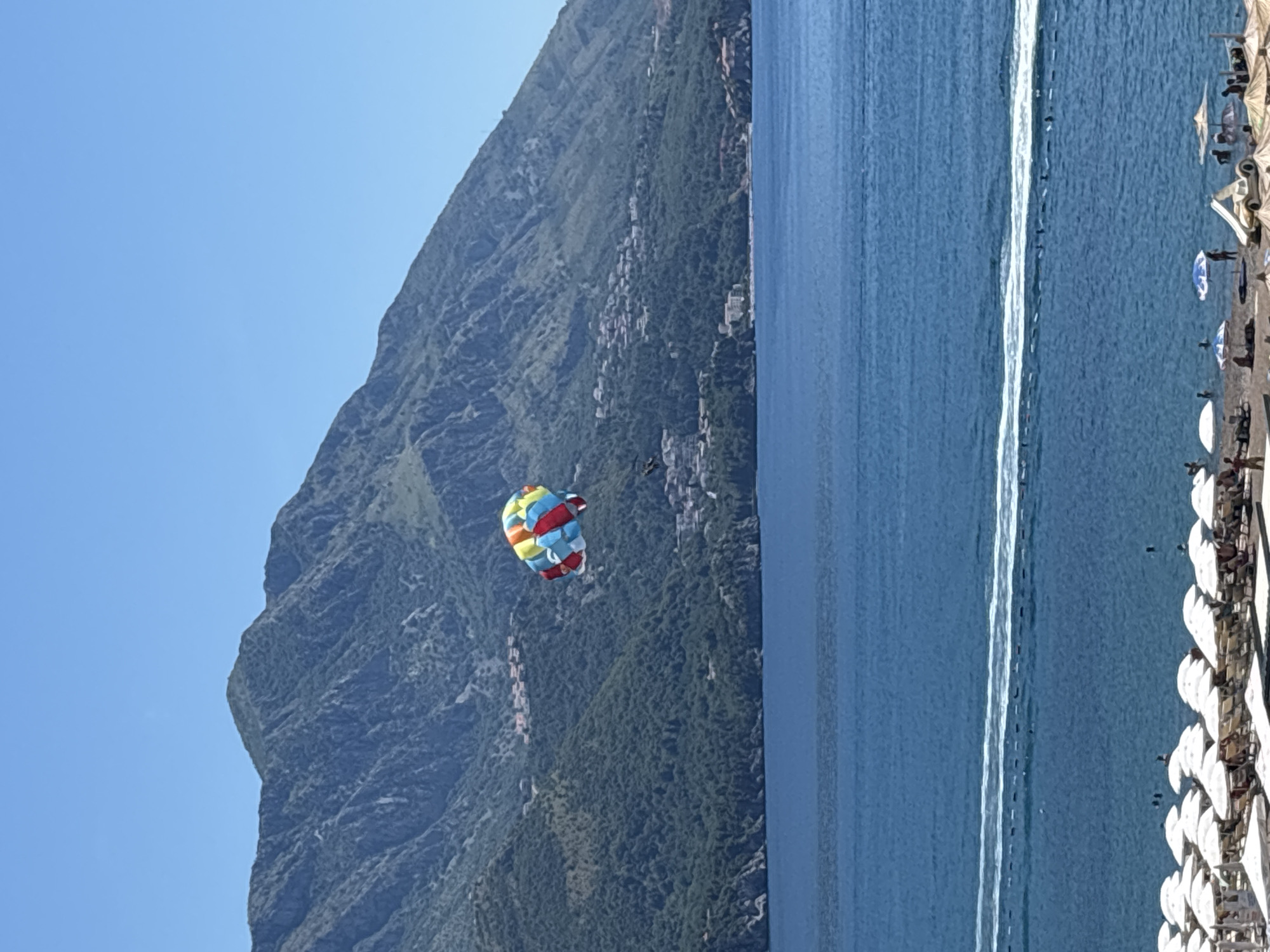 The image shows a wide shot of the coastline near Boreti, Montenegro on a clear, sunny day. In the foreground, a beach is populated with numerous white beach umbrellas, chairs, and a few people. The sea is a deep blue, with a white wake cutting across it, suggesting a boat is moving at speed. In the middle ground, a colorful parasail is visible in the sky, attached to a person being towed. Behind the sea and parasail, lush green hills rise steeply, dotted with small buildings and homes. The distant background is dominated by a large, rugged mountain range under a clear blue sky. The overall scene captures a popular tourist destination with recreational activities taking place.