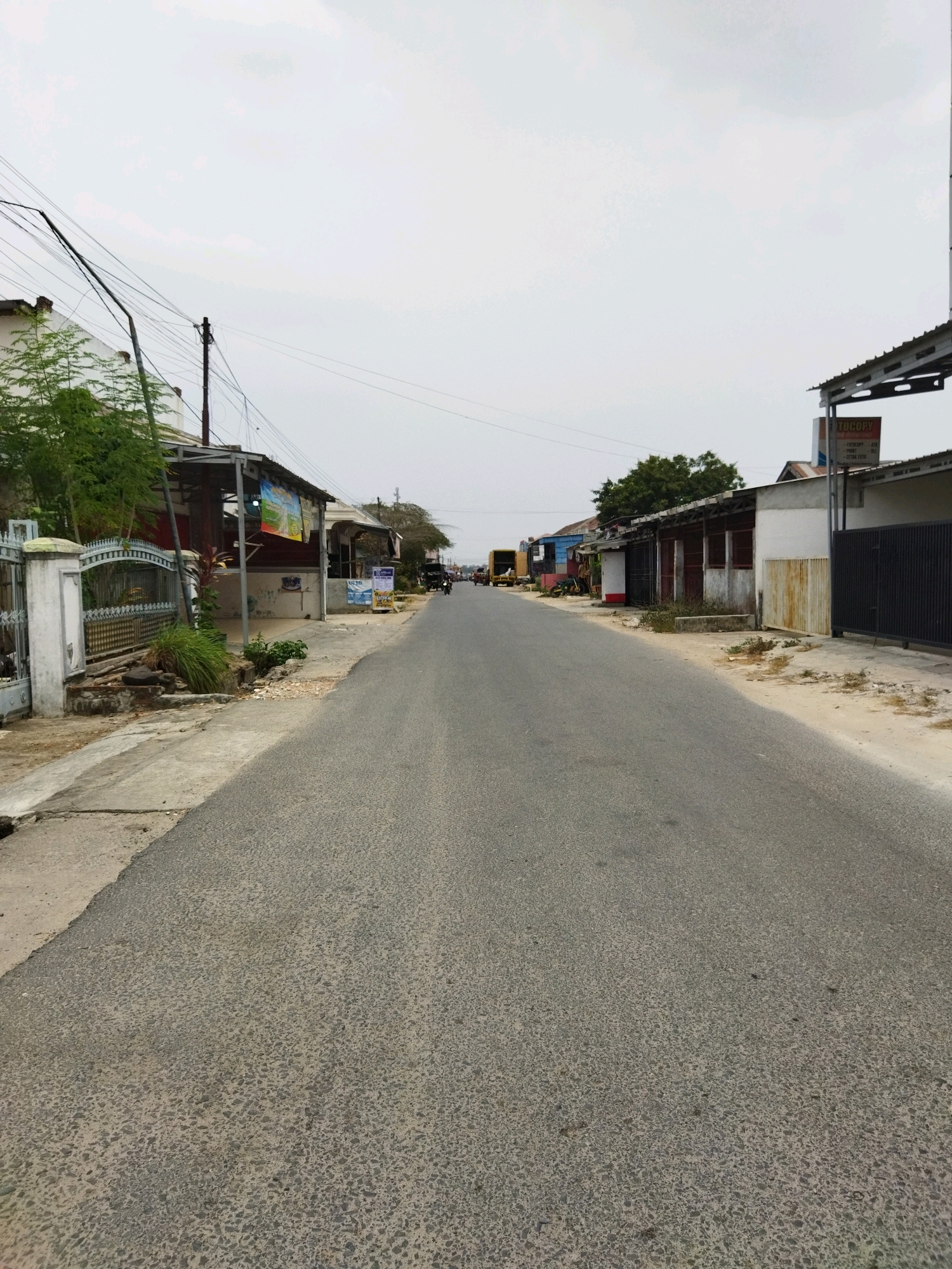 This is a vertical, eye-level, medium-shot photograph taken on a cloudy day on a paved road in Banjar Agung, Indonesia. The road stretches into the distance, lined on both sides by modest, single-story buildings and some trees. On the left side of the road, there is a light-colored fence and a building with a covered awning. A sign for "TOCOPY" is visible on a building on the right side of the road. There are a few scattered people and vehicles visible in the distance, suggesting moderate activity. The overall impression is of a simple, functional street in a rural or semi-urban area of Indonesia.