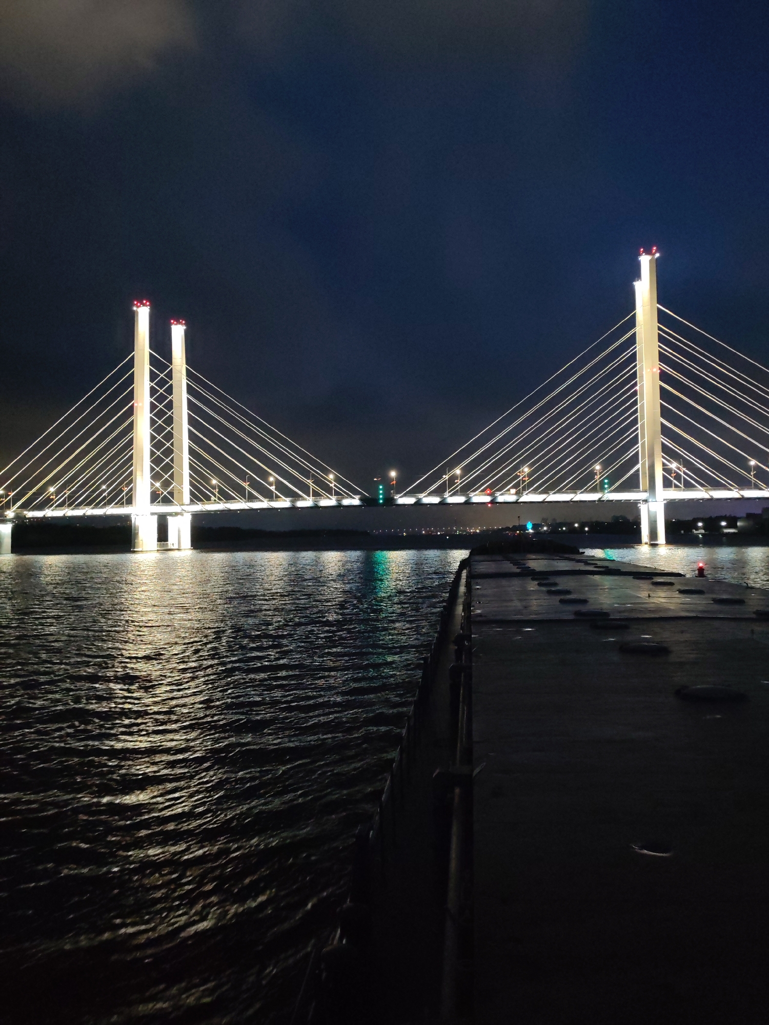 The image shows a bridge at night. The bridge is illuminated with white lights and has tall pillars with red lights on top. The bridge cables are arranged in a fan-like pattern. Below the bridge is a body of water with light reflecting on its surface. A dark structure, possibly a dock or pier, extends into the water from the foreground. The sky is dark with some clouds. The location is the city of Unknown, Unknown.