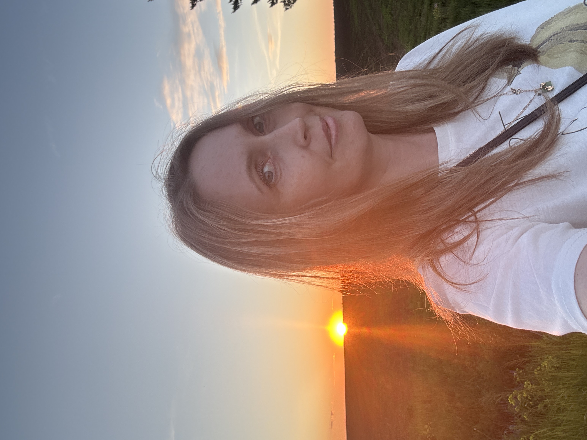 The image captures an adult female outdoors during sunset in Перевозное, Russia. She is positioned slightly to the right of center, looking directly at the camera with a subtle smile. Her long, light brown hair is parted on the left. The left side of her face and hair are bathed in direct, warm light from the setting sun. She wears a white short-sleeved top with a faint, light-colored graphic print on the chest, and a gold-toned necklace featuring multiple charms, including a dark green element. A dark strap crosses diagonally over her upper torso. In the background, the sun is partially visible on the left horizon, appearing as a bright orange semi-circle, emitting a distinct vertical light flare. The sky transitions from bright orange and yellow near the horizon to a clear light blue with sparse, wispy clouds higher up and to the right. The landscape behind her consists of expansive green fields with visible vegetation. On the far right, silhouetted tree branches are visible against the sky.