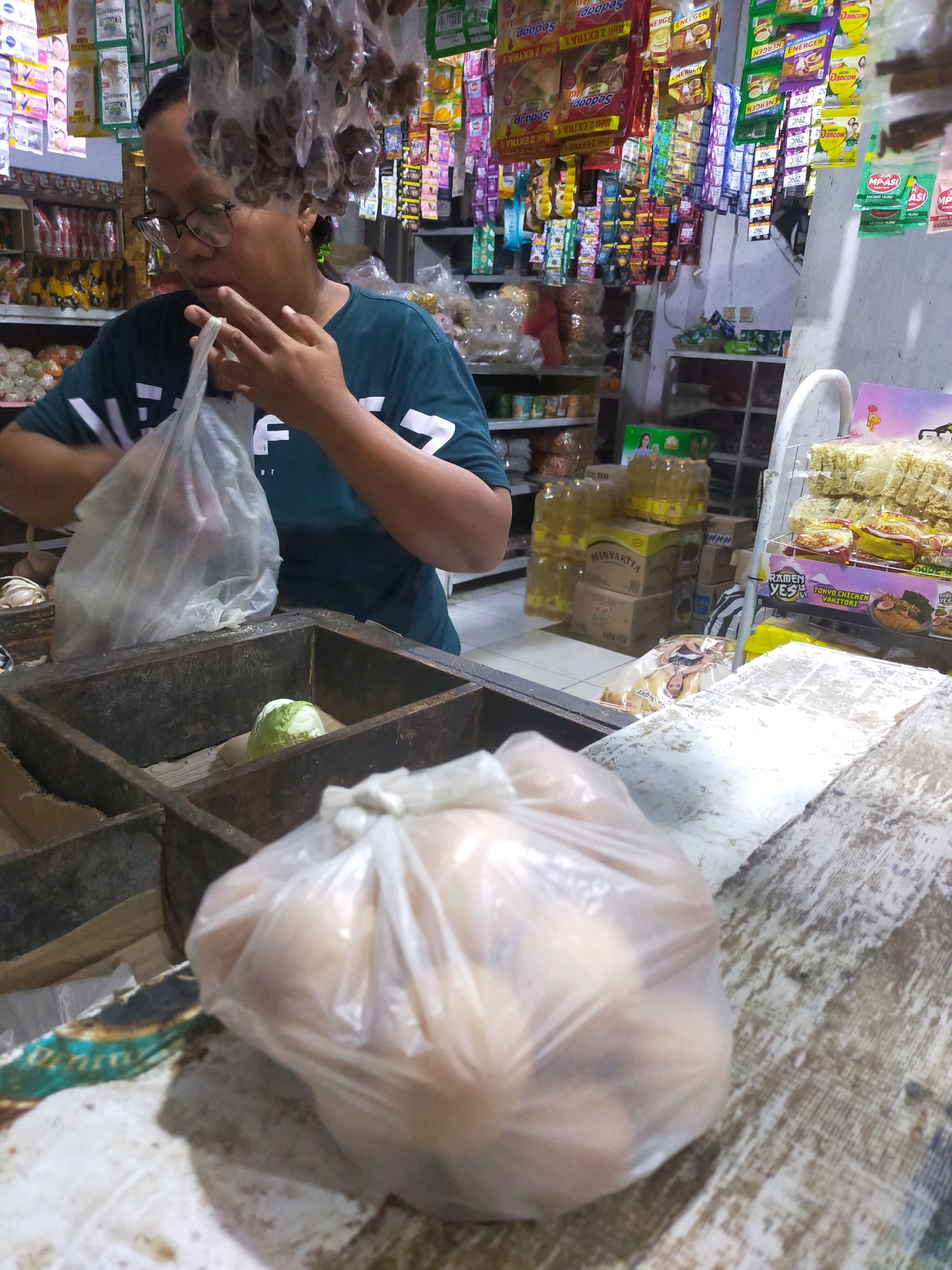 In Mojodanu, Indonesia, a person wearing glasses and a dark green t-shirt is standing in front of a wooden display case filled with various items. The person is holding a translucent plastic bag containing what appears to be food items. Inside the wooden display case, there is a head of lettuce in one section, and other items are visible in adjacent sections. In the foreground, another translucent plastic bag filled with round, light-colored objects, possibly eggs or potatoes, rests on a wooden surface. Behind the person, shelves are stocked with an array of packaged goods, including food products and beverages, indicating a retail environment such as a small shop or market stall. The overall scene depicts a transactional interaction or preparation for such in a local Indonesian setting.