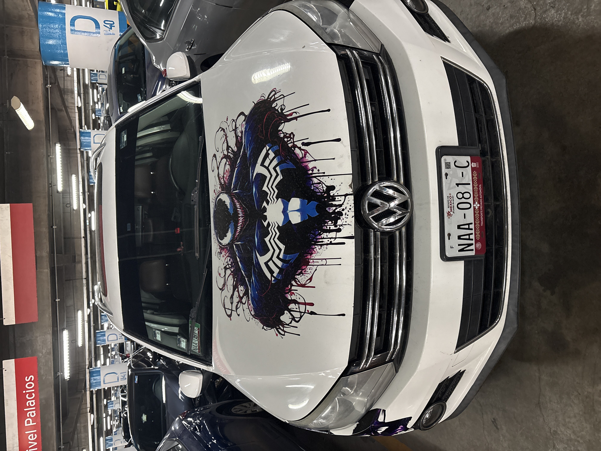The image shows a white Volkswagen car with a large Venom character graphic on its hood. The car is parked in an indoor parking garage, likely the "Nivel Palacios" area in Mexico City, Mexico, as indicated by signage. The license plate of the car reads "NAA-081-C" and it is registered as a "TRANSPORTE PRIVADO AUTOMÓVIL" in Mexico. Several other vehicles are parked nearby. The lighting is provided by overhead fluorescent lights.
