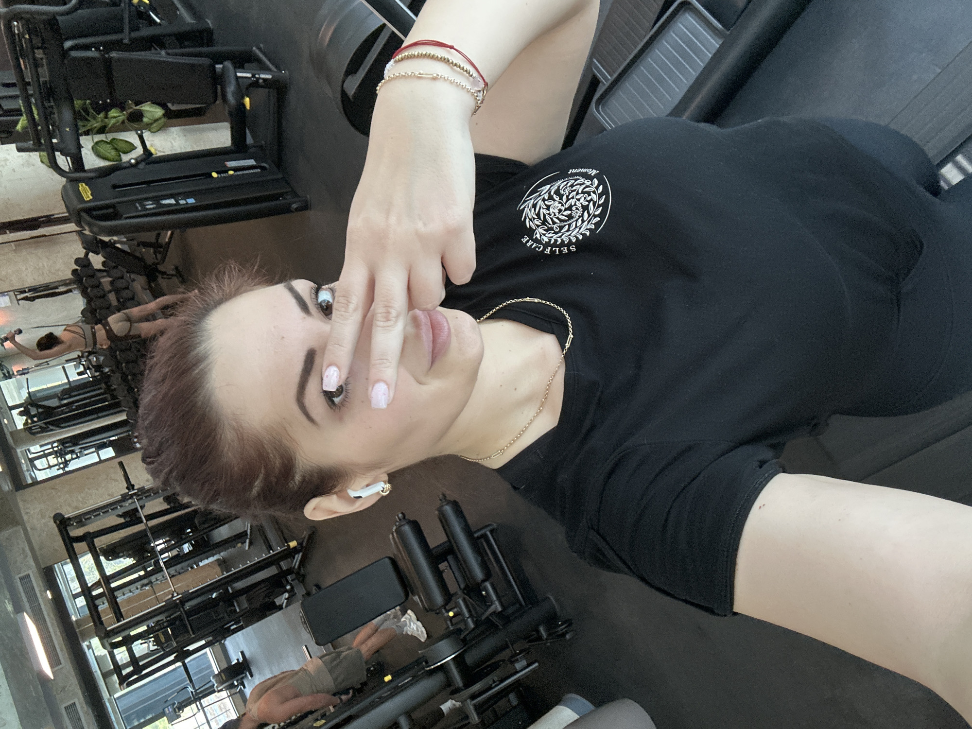 A woman poses for a selfie indoors at a gym. She wears a black T-shirt with a white "SELF CARE" logo, a gold necklace, and multiple bracelets on her left wrist. She is positioned in front of weightlifting equipment. Other individuals are visible in the background using the equipment. The location is Kazanlak, Bulgaria.