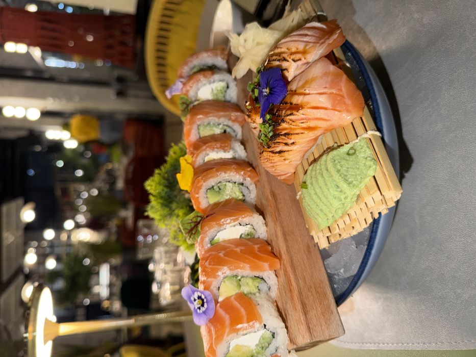 The image displays a sushi and sashimi platter presented on a table within a restaurant setting in Budva, Montenegro. In the foreground, a wooden serving board holds nine salmon, avocado, and cream cheese sushi rolls, garnished with small purple or yellow edible flowers and green leaves. To the right, on a bamboo mat situated atop crushed ice in a dark blue bowl, are two pieces of seared salmon nigiri, topped with a dark sauce, chopped chives, and a purple edible flower. Adjacent to the nigiri are portions of pickled ginger and a rectangular mound of wasabi on a separate bamboo mat. The platter rests on a light grey, textured surface. The blurred background indicates a dimly lit interior dining area, featuring numerous out-of-focus light sources, a tall lamp on the left, indistinct patrons including one wearing a yellow top, and various decorative elements such as a prominent red cylindrical object on the far right.