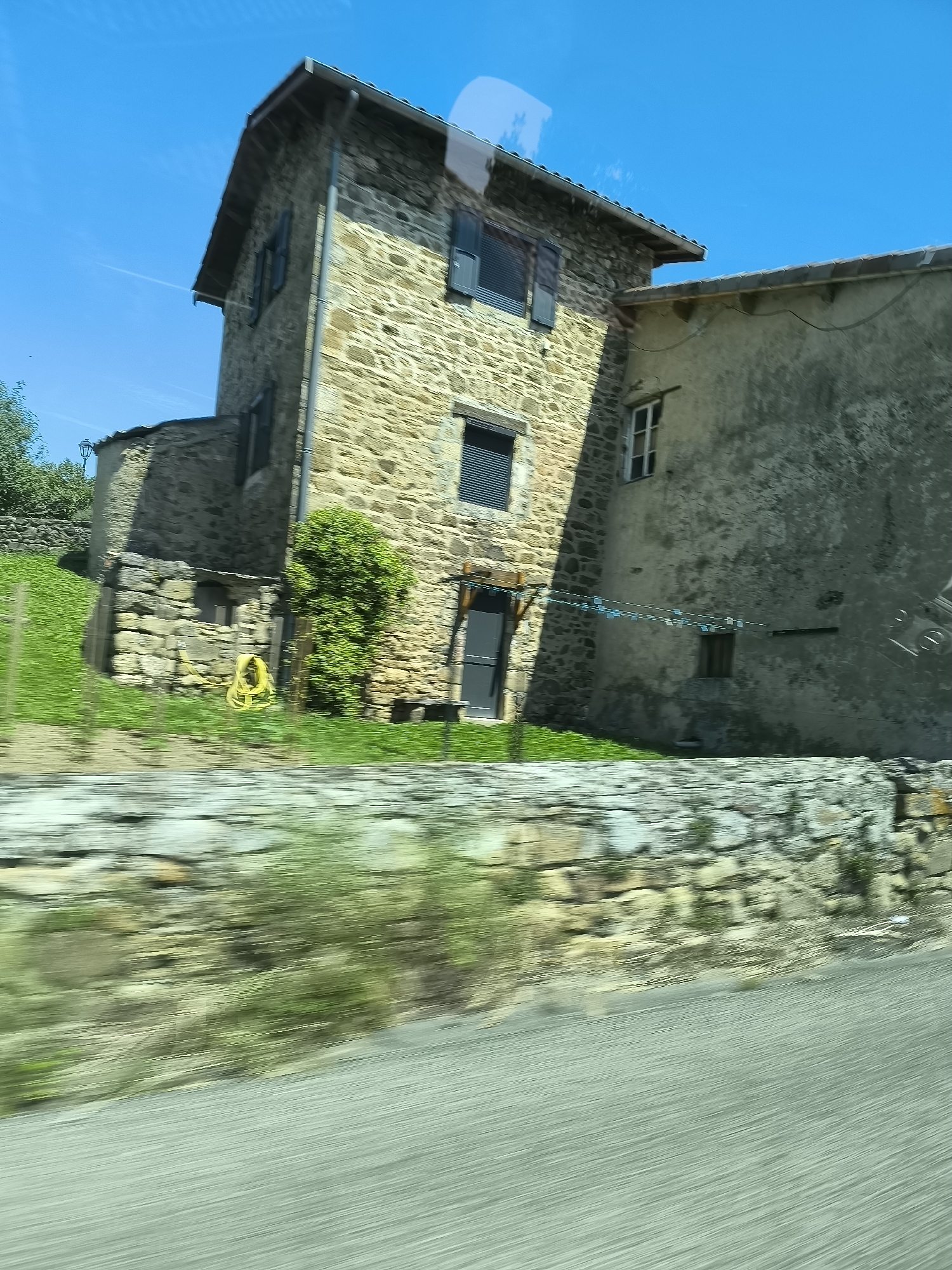 The image depicts a stone building complex in what appears to be a rural setting near Chavanay, France. The complex consists of a taller, stone-built section with a gable roof and dark window shutters, adjoined to a lower, plastered section. A stone wall runs along the lower edge of the frame, with green vegetation on the adjacent grassy slope. A yellow garden hose is visible next to a bush by the stone building. A clothesline hangs between the two buildings. The blurred foreground suggests the image was taken from a moving vehicle. The sky is clear and blue.