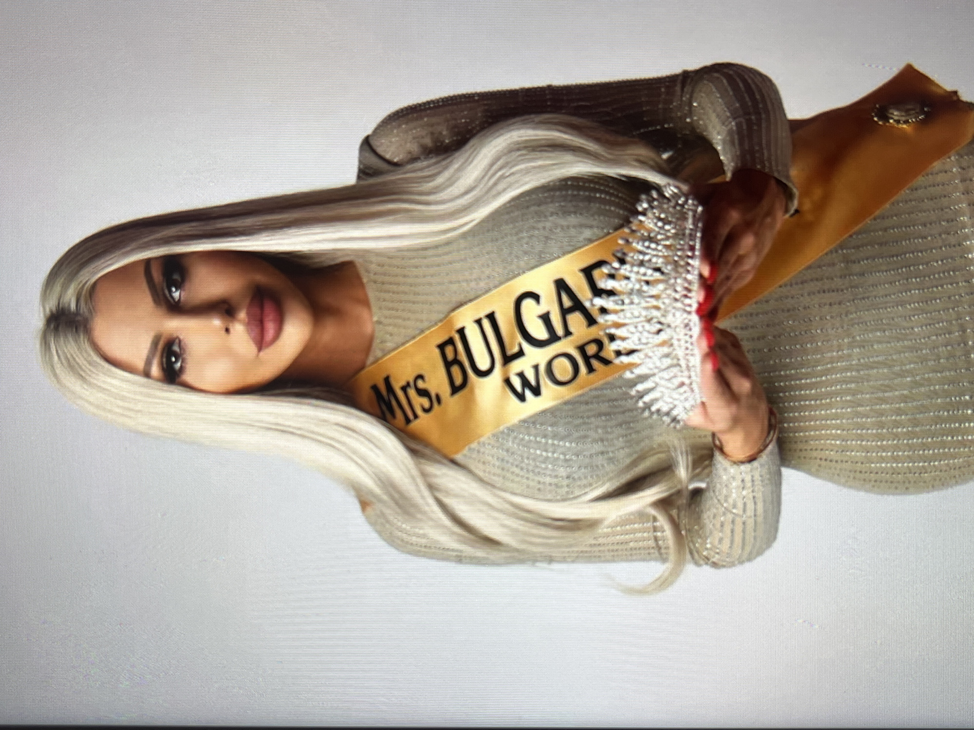 A woman is posing for a photograph. She is wearing a sparkly gold dress and a gold sash that reads "Mrs. Bulgaria World." She is also holding a silver crown. The background is plain white. The image appears to be taken indoors, likely in a studio setting. The lighting is bright and even. The woman has long blonde hair and is wearing full makeup. The overall mood of the image is celebratory, suggesting a pageant or similar event. There are no discernible time of day or weather cues. The location context of Vratsa, Bulgaria, is provided, but there are no visual cues in the image to confirm this.