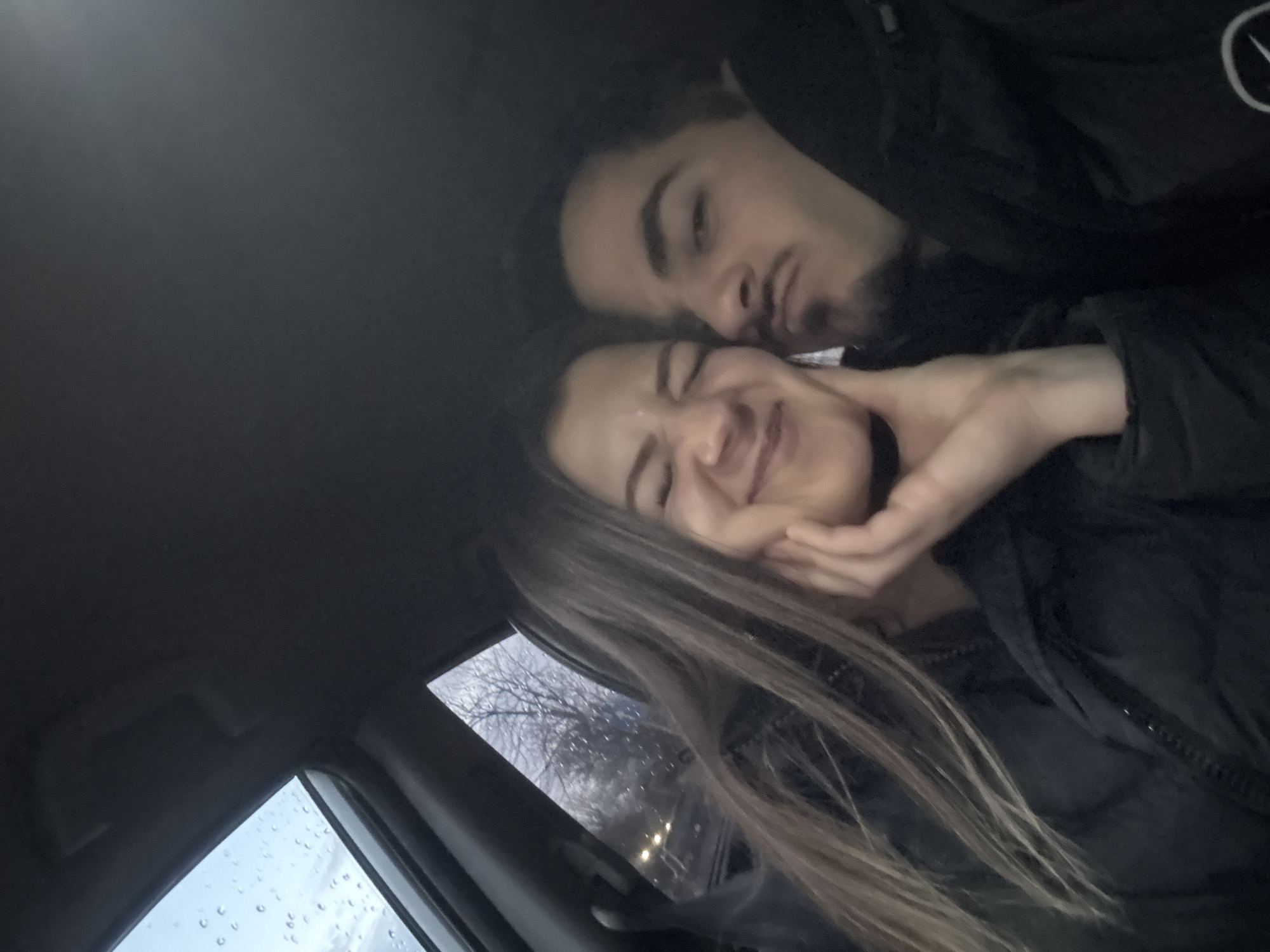 The image shows a man and a woman in a car. The man is leaning over the woman, appearing to kiss her cheek. The woman is smiling with her eyes closed, and the man has his hand on her face. It is raining outside. The location in the city of Sofia, Bulgaria cannot be confirmed from the image.