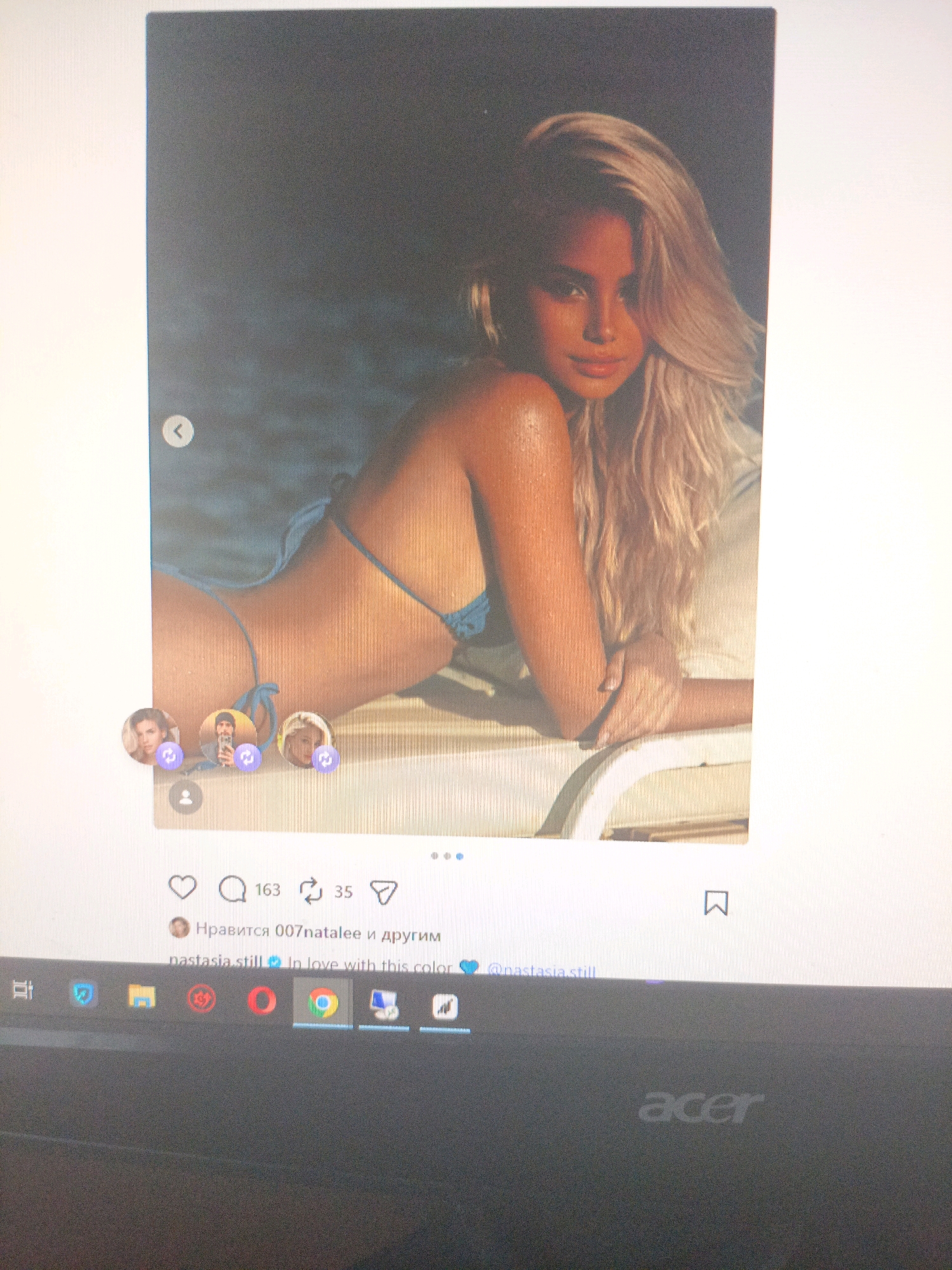 The image displays a screenshot of a social media post, likely from Instagram, viewed on a laptop screen. The post features a photograph of a woman with blonde hair wearing a blue bikini, posing outdoors. The background appears to be a beach or poolside scene with water and sand. Below the photograph, there are interactive elements common to social media platforms, including likes (indicated by a heart icon and the number 163), comments (indicated by a speech bubble icon and the number 35), and a share icon. A caption below these icons reads "Нравится 007natalee и другим" which translates to "Liked by 007natalee and others." Further text reads "nastasia.still In love with this color". The screenshot also shows the laptop's taskbar at the bottom, with application icons like Google Chrome. The setting is indoors, as evidenced by the laptop screen. There is no visible information within the image to indicate the location in the city of Ухманы, Russia.