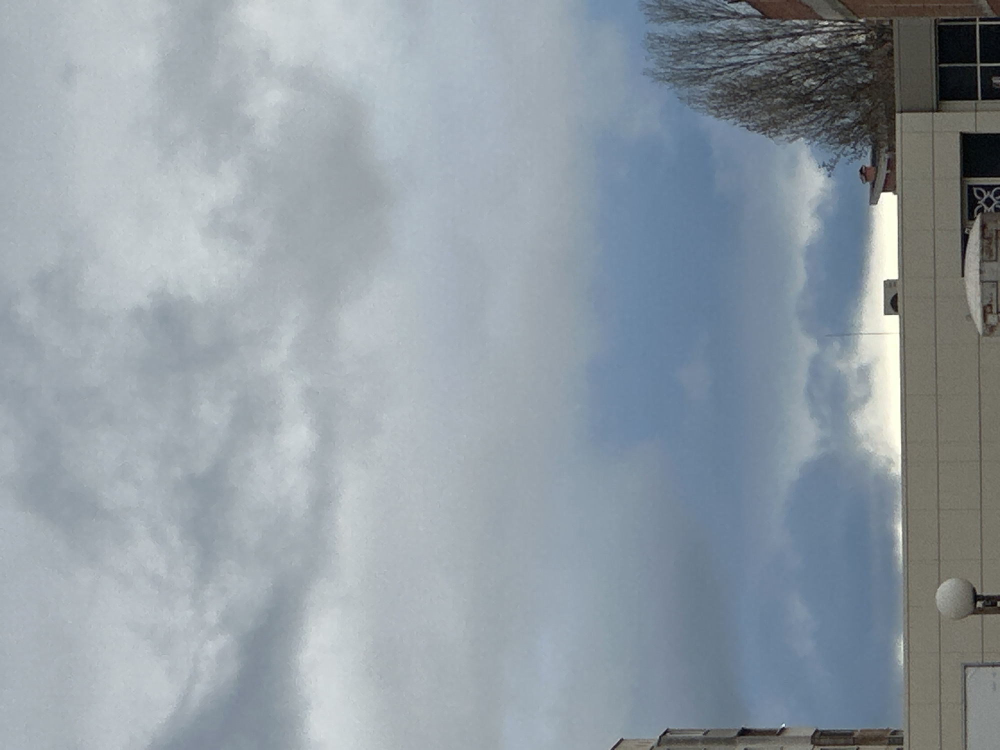 The image shows a view of the sky, partially obscured by clouds, alongside the corner of a building. A bare tree branch extends into the upper right corner. The building is a light color, with rectangular windows and architectural details such as a streetlamp and an air conditioning unit. Parts of other buildings are visible. Based on the buildings and architectural style, it is possible that the location is Sofia, Bulgaria.