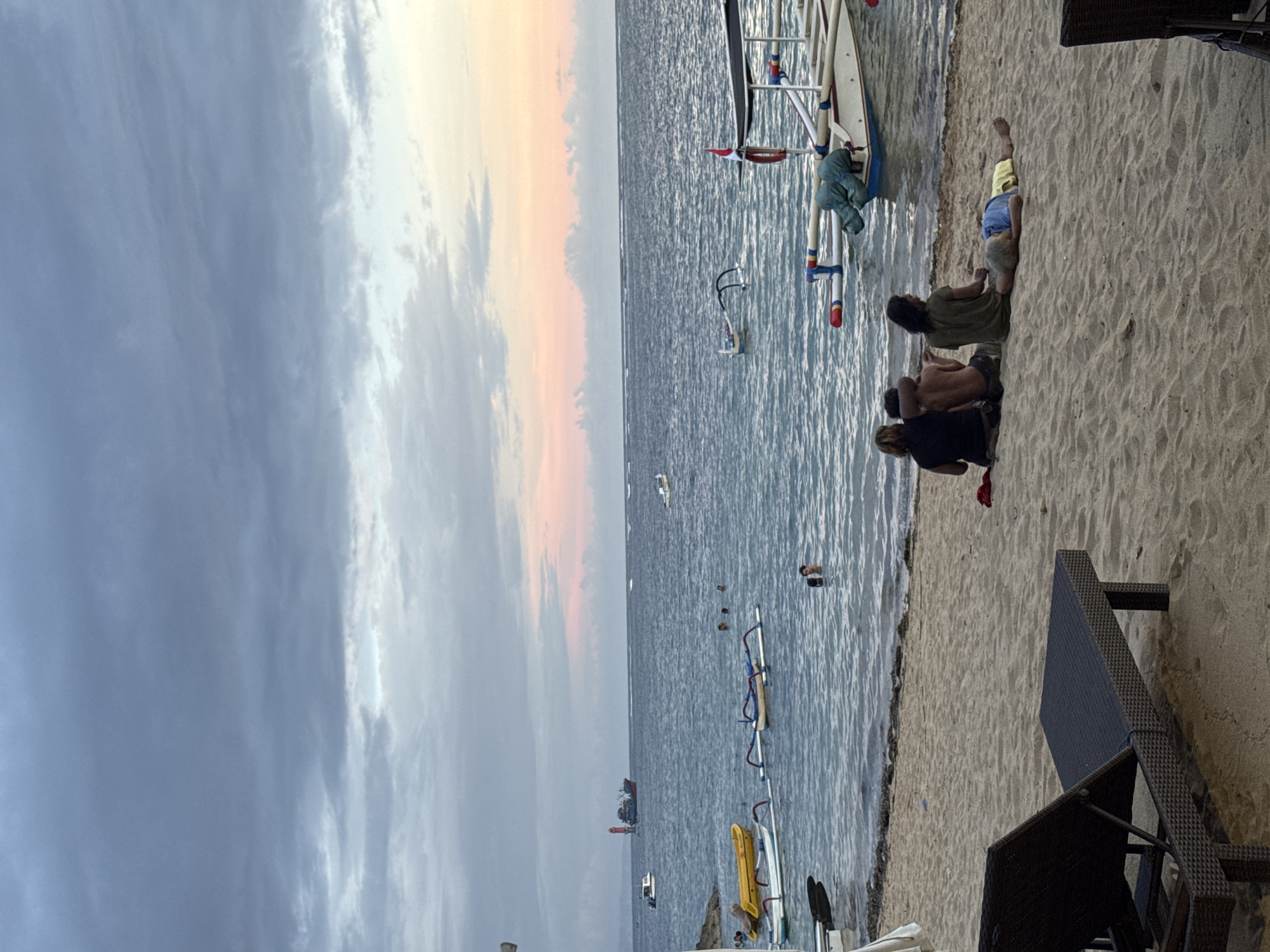 The image depicts a beach scene in Denpasar, Indonesia. The sky is cloudy with hints of a sunset. The ocean is calm with several boats and what appear to be windsurfing rigs in the water. Along the shoreline, a small group of people are relaxing on the sand, including one person lying down. A boat is partially beached. Beach chairs are visible.