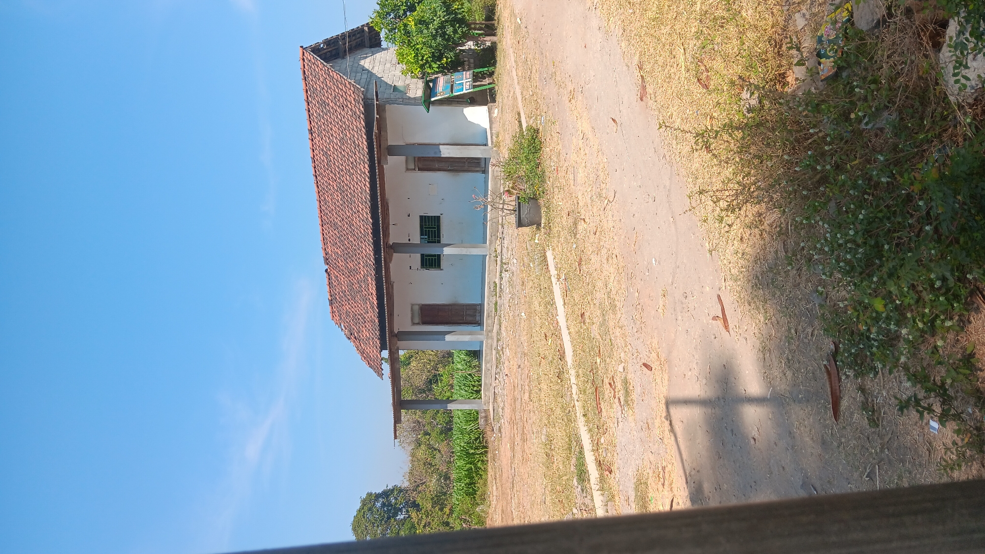 A wide shot of a single-story, white building with a red tile roof under a clear blue sky in Gemenggeng, Indonesia. The building has a covered porch supported by three gray pillars, with two wooden doors and two windows with green bars on its front facade. A small, potted plant sits in front of the building, and to the right, a green signpost stands near a leafy tree. The foreground shows a dirt path and dry grass.