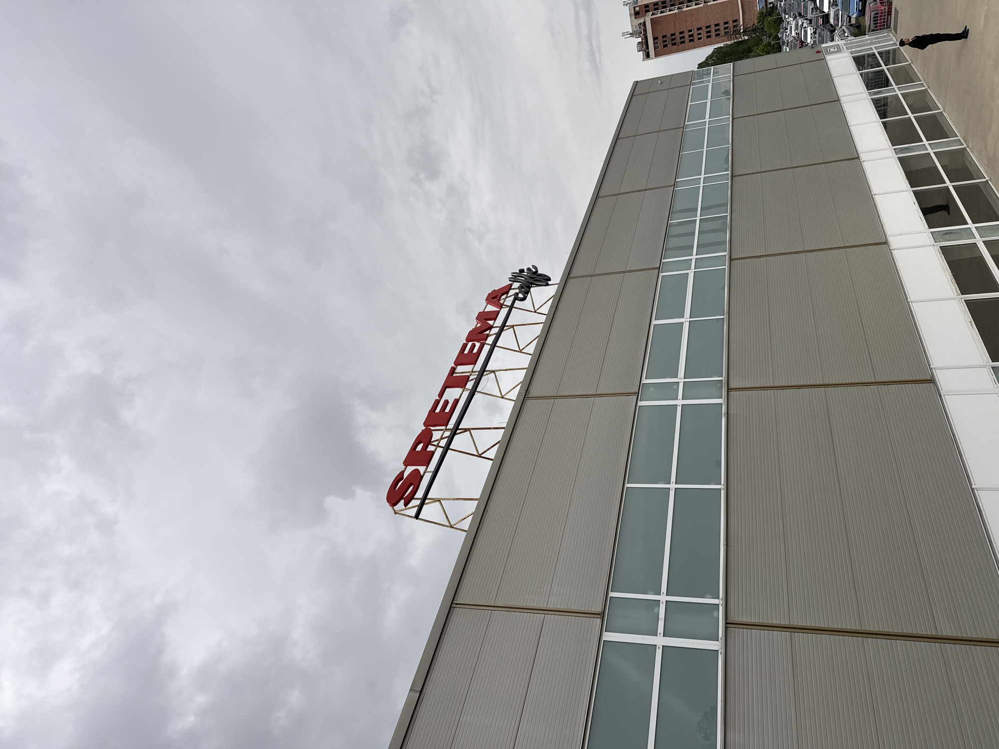 The image shows an exterior shot of a building. The sign on top of the building reads "SFETEM". There is a man standing in the upper right corner. The sky is overcast with clouds. The building has a gray facade with vertical panels and a section of windows. In the background, a tall brick building can be seen. This photo was taken in Sofia, Bulgaria.