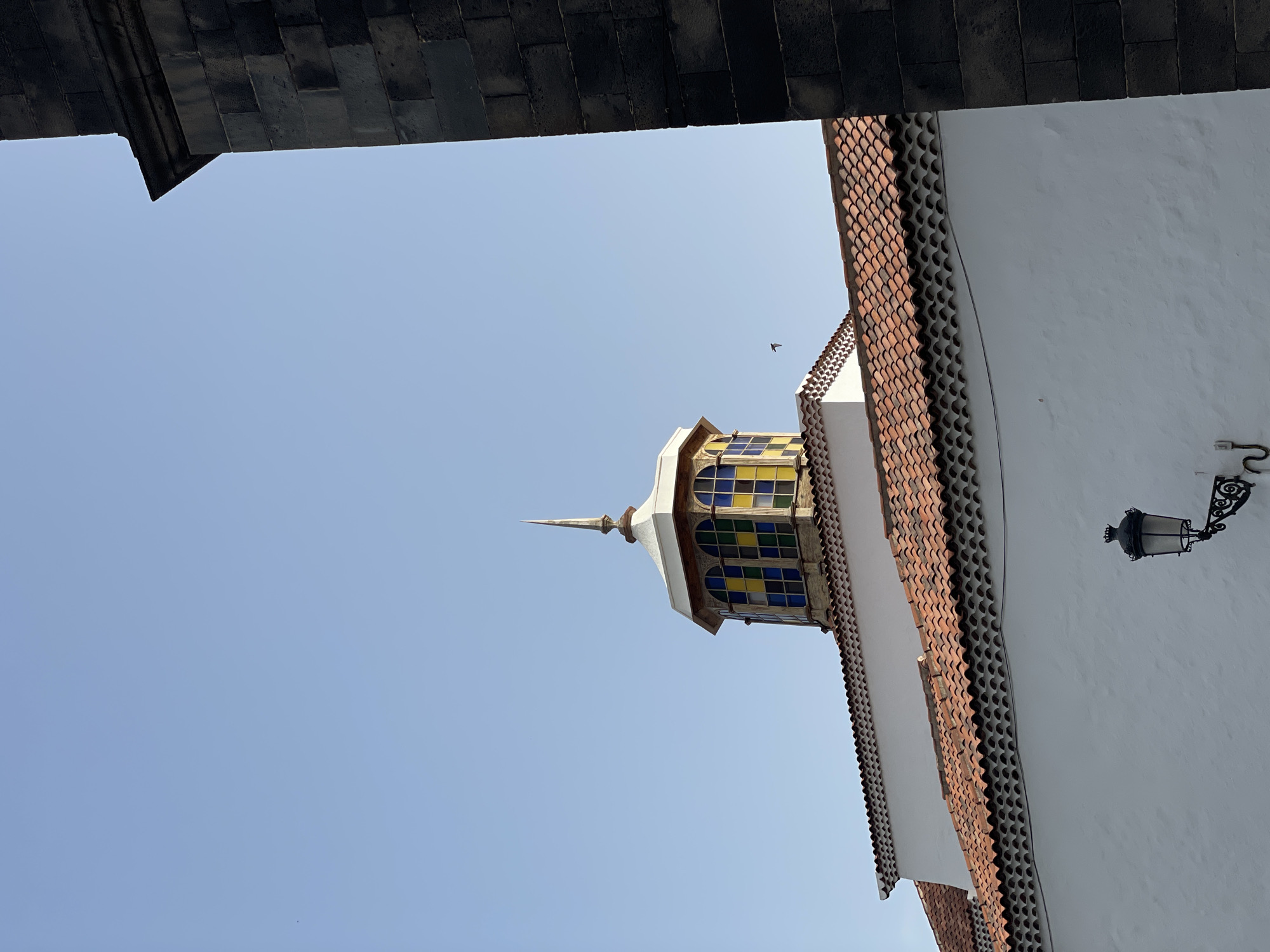The photograph shows a partial view of the Iglesia de la Concepción in Santa Cruz de Tenerife, Spain, framed by a dark stone structure above. The church's steeple, featuring a colored glass window pattern of yellow and blue, extends upwards into a clear blue sky. A white building facade is visible to the right, adorned with a reddish-brown tiled roof and a black ornate lamppost. A bird is seen flying in the sky above the steeple.