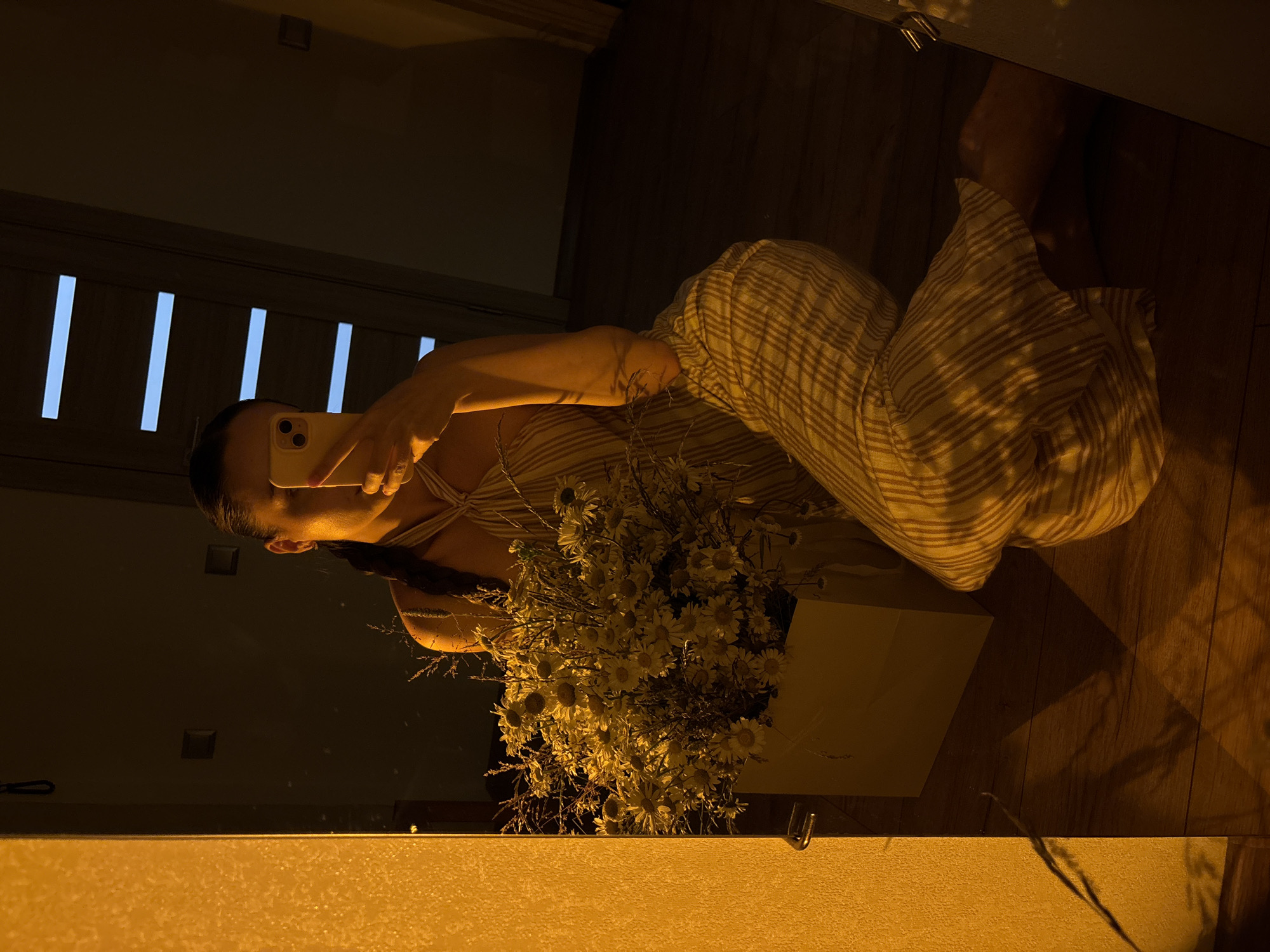 A person is kneeling on a wooden floor in front of a mirror, taking a selfie with a smartphone. The person is wearing a striped jumpsuit and has a braid that extends down their back. To their left, a large bouquet of daisies is contained within a white box. The room is dimly lit with warm, yellow light, casting shadows on the floor and walls. The mirror reflects the person, the bouquet, and a doorway with horizontal light blue accents. The overall ambiance is intimate and personal.