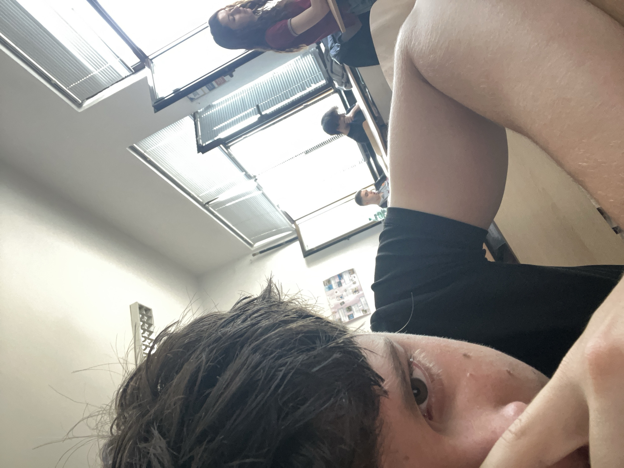This media file captures an upward-looking, rotated perspective, as if the photographer is lying down or seated low, gazing towards the ceiling. In the immediate foreground, the right side of a person's face is visible, featuring a dark brown eye, part of their nose, and a hand partially covering their mouth. Their messy, dark brown hair frames the left side of the shot, and they appear to be wearing a black top. A bare leg or arm, showing fine body hair, occupies the upper right portion of the frame.
The background reveals an indoor environment, likely a classroom, office, or communal study area, illuminated by several rows of bright fluorescent lights recessed into a white ceiling. Due to the camera's orientation, the ceiling appears as a bright, dominant surface. Several other individuals are visible in the mid-ground and background, appearing partially rotated or upside down. One person with long, wavy dark hair and a red top is seated on the left side, seemingly at a desk or table. Further back, two more individuals can be seen; one has dark hair and is looking downwards, while another is even further in the distance.
The activity suggests a group of people occupying a room, possibly engaged in studying or working, given the interior setup. No specific actions are clearly discernible. There is no legible text visible within the frame. The time of day and external weather conditions cannot be determined from this indoor scene. The provided context places the location in Zlatograd, Bulgaria.