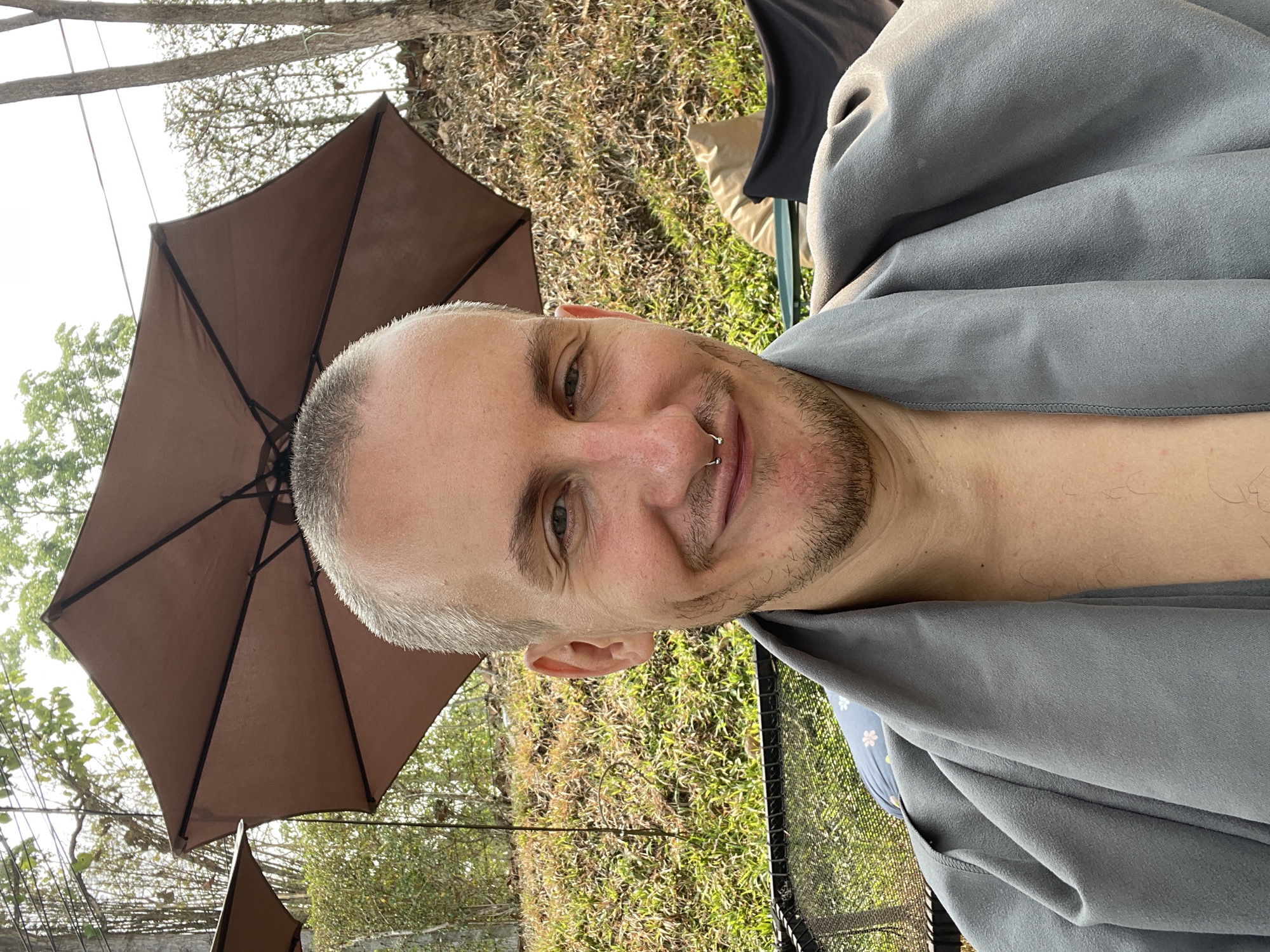 A man with short, light gray hair, a nose ring, and stubble is positioned in front of a brown patio umbrella. He is wearing a gray garment, and his chest is exposed. The setting appears to be outdoors, with vegetation and another umbrella visible in the background. The photo was taken in Tambon Nam Phrae, Thailand.