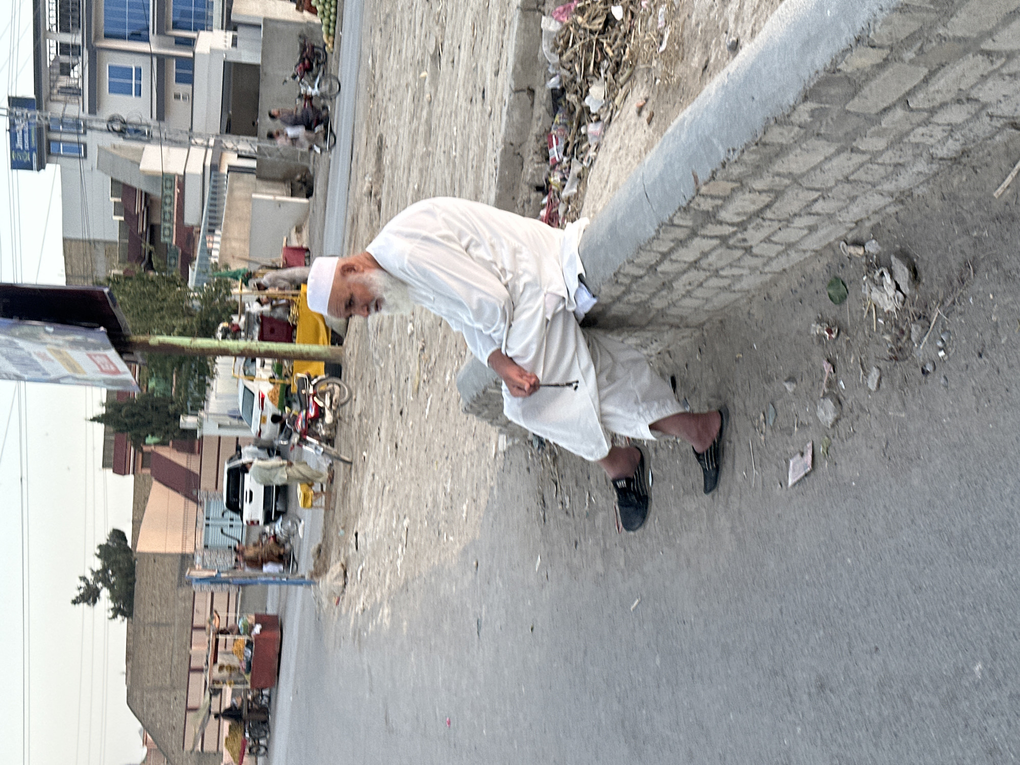 An elderly man with a white beard and a white cap sits on a low brick wall beside a street in Quetta City Tehsil, Pakistan. He is wearing a white shalwar kameez and black sandals, and is holding a string of prayer beads. The street is paved, and there are shops and buildings in the background. There is also some trash on the side of the road. The man appears to be relaxed, and is looking off to the side. The lighting suggests it is late afternoon or early evening.