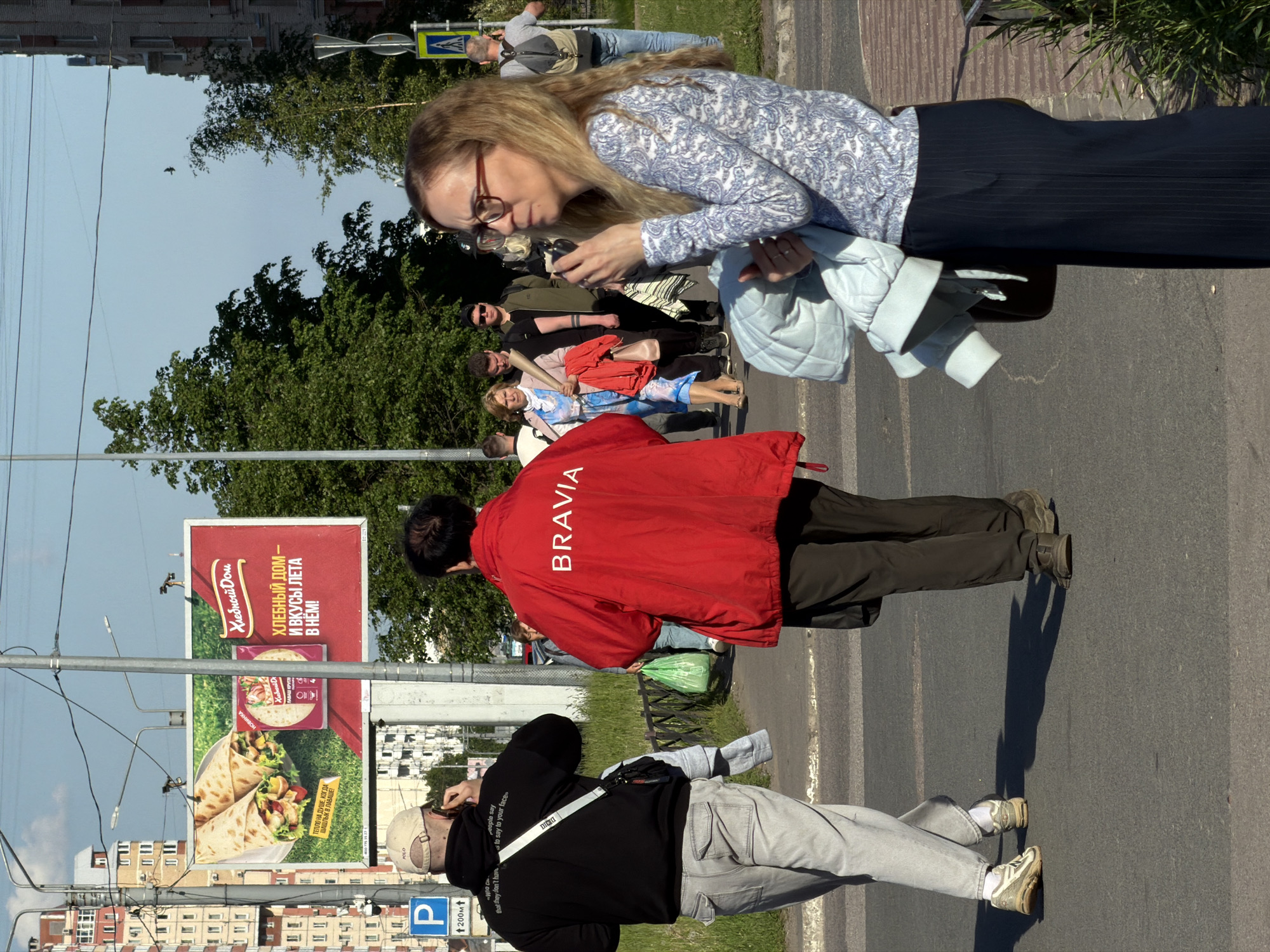 The image captures an outdoor street scene in Saint Petersburg, Russia, on a clear, sunny day, as evidenced by distinct shadows. Several pedestrians are visible on what appears to be a sidewalk or street.
In the foreground, a person is seen from behind, wearing a black hoodie with partial text including "People say," "to say your face," and "that they don't have." They wear light grey pants and a light-colored cap with "POLO" visible, and appear to be interacting with a phone. To their right, another individual is viewed from behind, dressed in a bright red jacket prominently displaying "BRAVIA" in white capital letters on the back, dark pants, and shoes.
Further back, a woman with long light brown hair and glasses is engrossed in her phone. She wears a light-colored patterned long-sleeved top and dark pants/skirt, carrying a light blue quilted jacket. Behind her, other pedestrians are partially visible, including one in a light blue patterned outfit and another in red/orange.
The background features residential buildings, lush green trees, and utility poles with overhead wires. A prominent billboard displays Russian text: "Хлебный Дом" (Bread House) with the slogan "И ВКУСЫ ЛЕТА В НЕМ!" (AND SUMMER FLAVORS IN IT!). It advertises food wraps/shawarma with additional text "ЛАВАШ КРУГЛ" (ROUND LAVASH) and "ТЕПЛО НА ДУШЕ, КОГДА ШАШЛЫК В ЛАВАШЕ" (WARM IN THE SOUL, WHEN SHASHLIK IN LAVASH). A blue 'P' parking sign indicates "200м" (200 meters), and a crosswalk sign is also present. The individuals are casually dressed, seemingly engaged in daily activities.