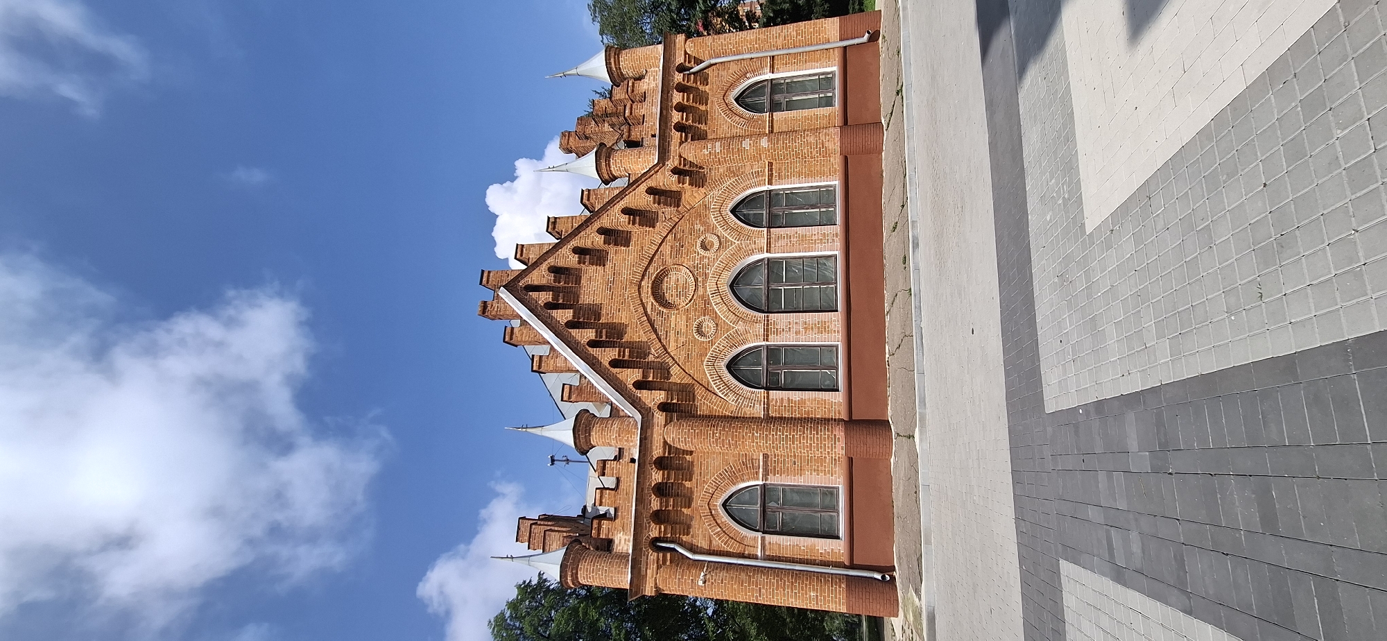 An exterior, eye-level, vertical shot shows a historic, red-brick building with arched windows and Gothic architectural elements. The building features decorative crenellations, pointed turrets, and brickwork detailing above the windows. A downspout is visible on the left side of the building. The building is situated on a paved area with light and dark grey rectangular pavers. Trees are visible on the left and right sides of the building, and the sky above is blue with scattered white clouds. The building is located at Amur, 17 Kommunisticheskaya Street, Blagoveshchensk, Russia.