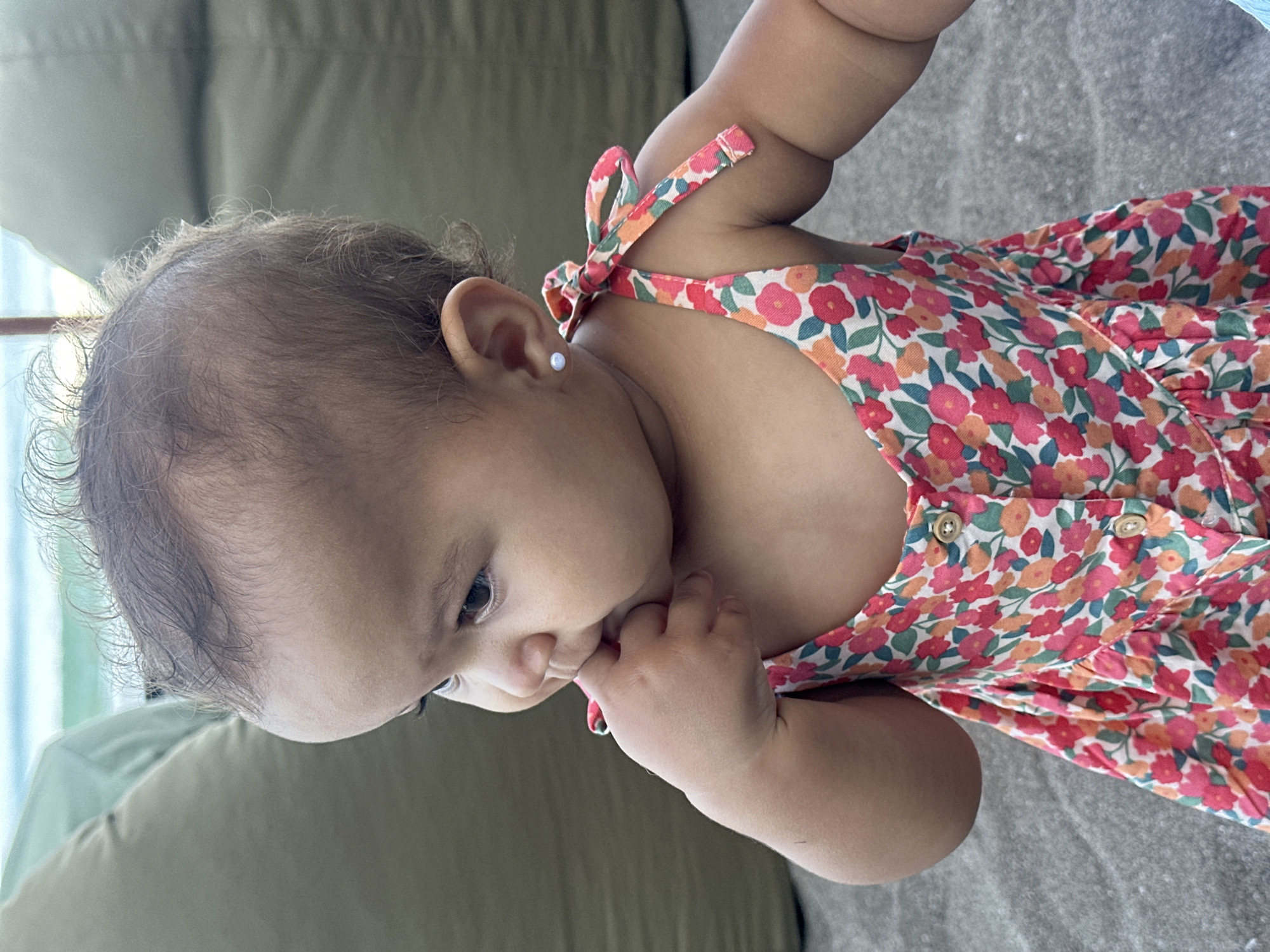 A baby is captured in a close-up, side-profile shot, angled downwards and to the left. The subject is a young child with dark, fine, curly hair, seen looking intently downwards. Their medium skin tone is visible, and a small, light-colored stud earring is present in their left ear.
The baby is wearing a vibrant floral dress or romper, patterned with numerous small flowers in shades of pink, orange, and red, accented with green leaves on a light background. The garment features shoulder straps tied in bows and two visible circular buttons on the front. One of the baby's hands is brought up to their mouth, with fingers possibly in their mouth or pressed against their cheek, a common action for infants.
The background is softly blurred, showing muted tones of green and grey, suggesting an indoor setting, possibly on or near upholstered furniture like a sofa. Bright, natural light illuminates the scene from the left, indicating it is daytime. The baby appears calm and focused on something out of frame below them. There is no visible text in the image.