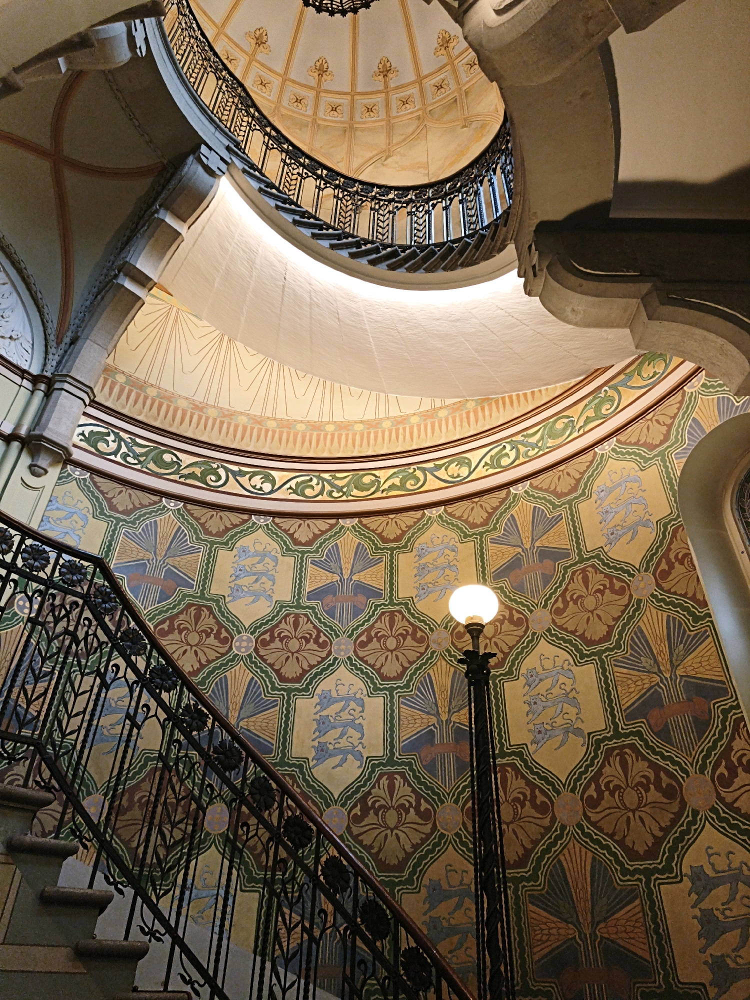 The image shows a view looking up at a grand, circular staircase with ornate metal railings and detailed murals on the curved wall. The staircase is made of stone steps and the railings are black, featuring intricate scrollwork and floral motifs. Above, the ceiling is a domed structure with decorative patterns and a large central opening allowing light to filter in.
The wall is covered in a vibrant mural with a repeating geometric pattern. Within these geometric shapes, there are stylized depictions of heraldic lions, floral designs, and what appear to be sheafs of wheat. The colors used in the mural are primarily yellow, green, brown, and blue.
A black lamp post with a spherical globe light stands near the staircase, illuminating the area. The overall architecture suggests a historical and impressive building. This scene is located within the Kiek in de Kök Museum in Tallinn, Estonia, specifically in the Great Coastal Gate.