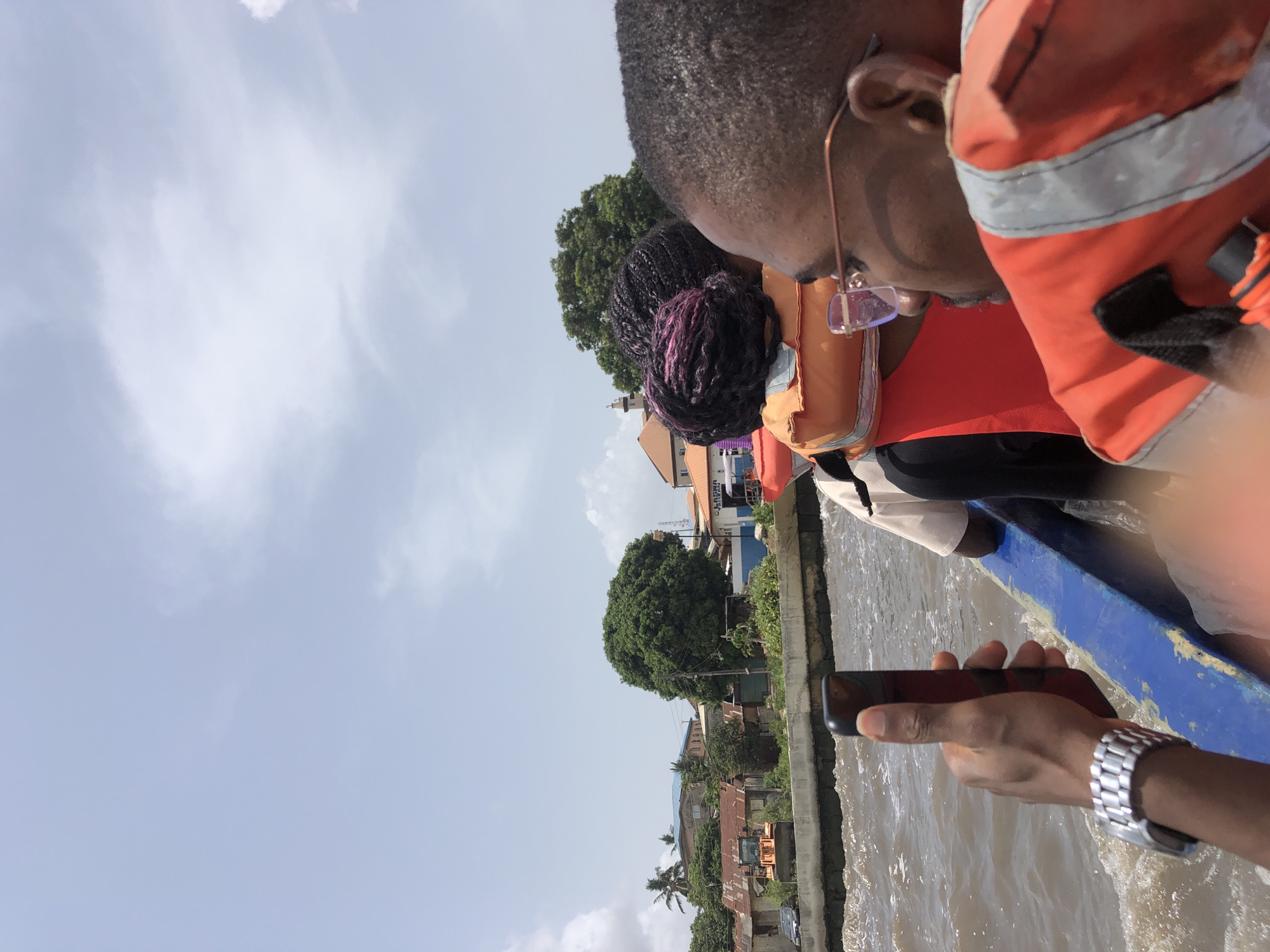 This image captures a boat journey on a sunny day, likely on a river or canal. In the foreground, on the right side of the frame, several individuals are seated in a blue boat, predominantly viewed from behind or the side. The closest person wears a prominent rust-orange life vest over a red shirt, has dark, short hair, and spectacles. Behind them, another individual with braided dark hair, featuring noticeable purple streaks, also wears an orange life vest. In the lower right corner, a hand wearing a silver watch holds a black smartphone, suggesting the person is capturing the scene.
The boat is positioned on a body of water with visible ripples and reflections. The opposite bank, on the left side, showcases a vibrant landscape featuring numerous buildings and houses, indicating a populated waterside community. Abundant green foliage, including large trees and distinct palm trees, is interspersed among the structures. The sky overhead is bright and mostly clear, with a few wispy white clouds, confirming daytime and fair weather conditions. On one of the buildings in the background, faint partial text, possibly "LATINIKA" and "ATERIAL," is visible. The scene conveys a sense of travel or exploration within a bustling, verdant environment.