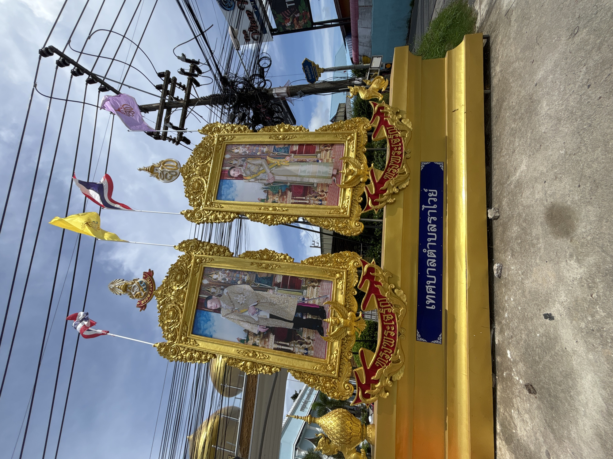 In Rawai, Thailand, an outdoor scene features two ornate golden frames displaying portraits of a king and queen. The frames are mounted on a gilded tiered platform. Thai flags, including the national flag and a purple flag with Thai characters, fly above the portraits, attached to poles and a utility pole. Electrical wires are visible in the background, along with a blue sign with white Thai lettering and the golden inscription "ทรงพระเจริญ" (Long Live the King) on red banners beneath the portraits. The ground is paved with concrete, and a grassy slope is present to the right.