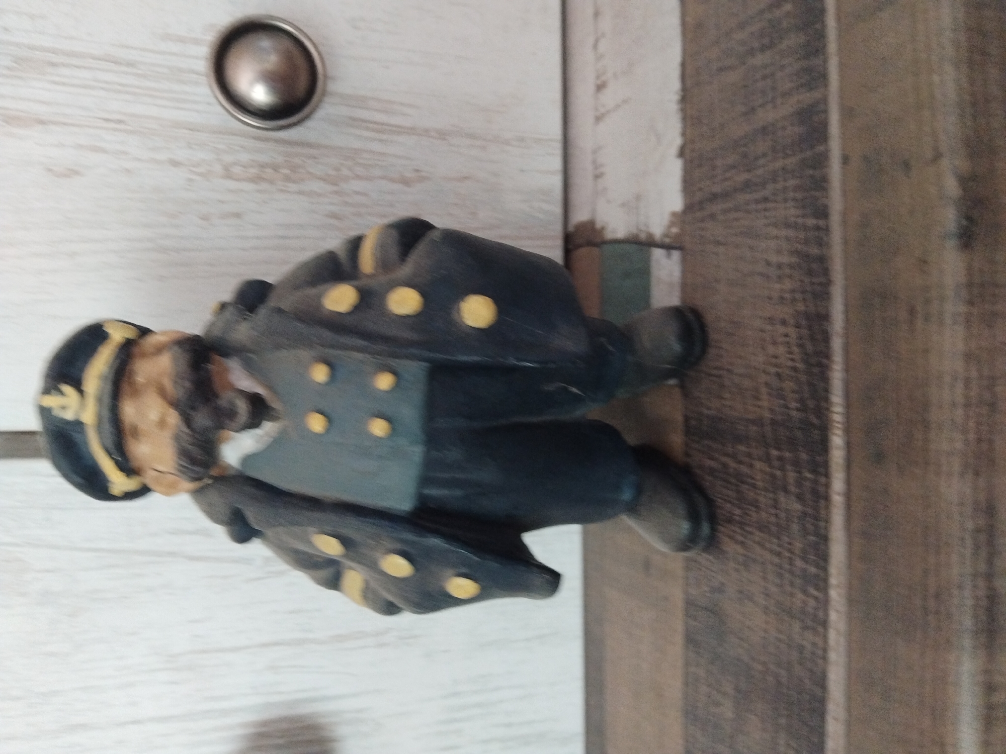 This is a full-shot, eye-level photograph of a figurine of a man dressed as a ship captain. The figurine stands upright on a wooden surface. The captain wears a dark blue uniform with gold buttons, a dark hat with a gold anchor emblem, and a brown mustache. The background consists of a white, vertically paneled wall with a round, silver decorative knob to the right of the figurine. The floor beneath the figurine is a wooden plank surface with a distressed, weathered appearance. The lighting is neutral, casting no significant shadows. The photograph was taken indoors, with no discernible external environmental factors. There are no people or animals present in the image, only the solitary figurine. The context of this image does not provide any information about its location in the city of Kyiv, Ukraine.