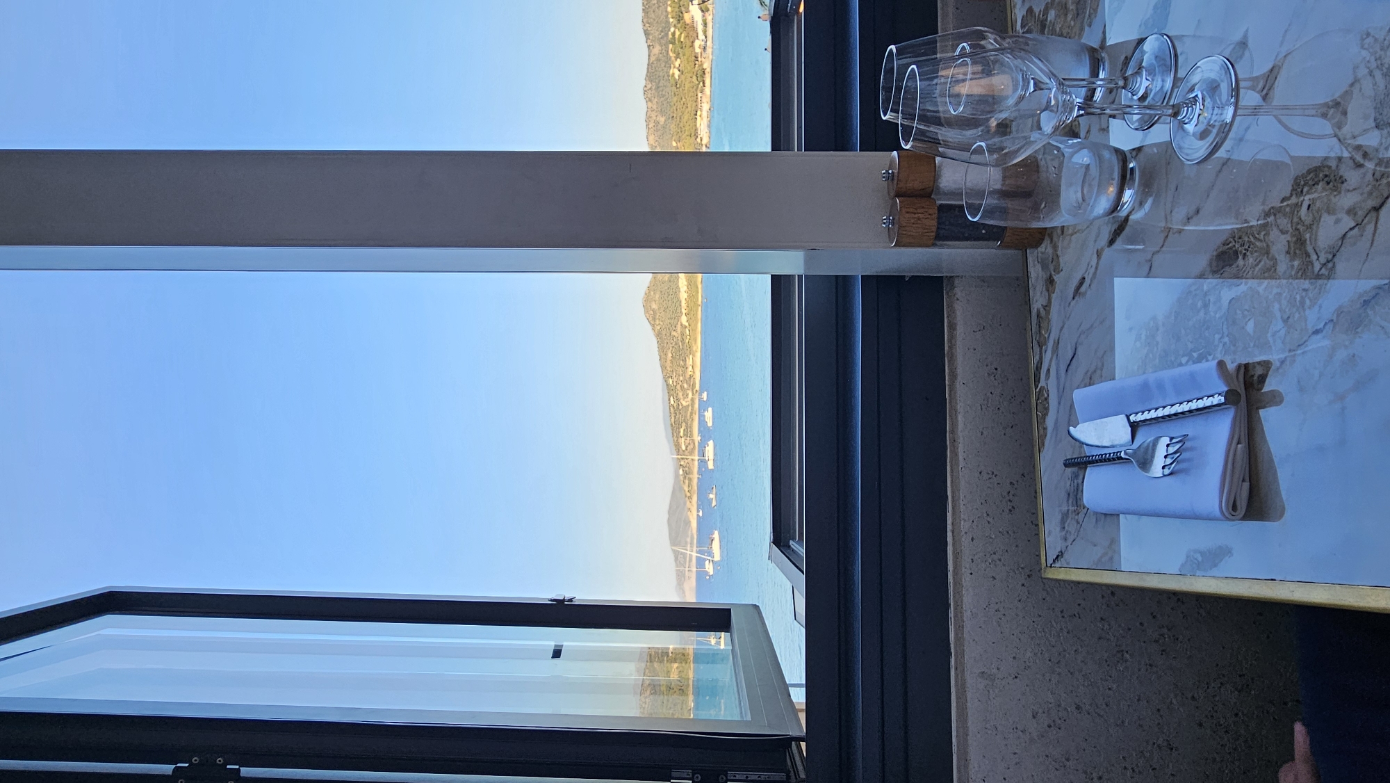 The image shows a table setting with cutlery and wine glasses arranged on a marble-topped table, viewed through an open window. Outside the window, the blue sea is dotted with sailboats, and a coastal landscape with hills and vegetation stretches into the distance under a clear blue sky. The setting suggests a restaurant with a scenic view, possibly in L'Île-Rousse, France, given the Mediterranean coastline. The sunlight indicates it's daytime.