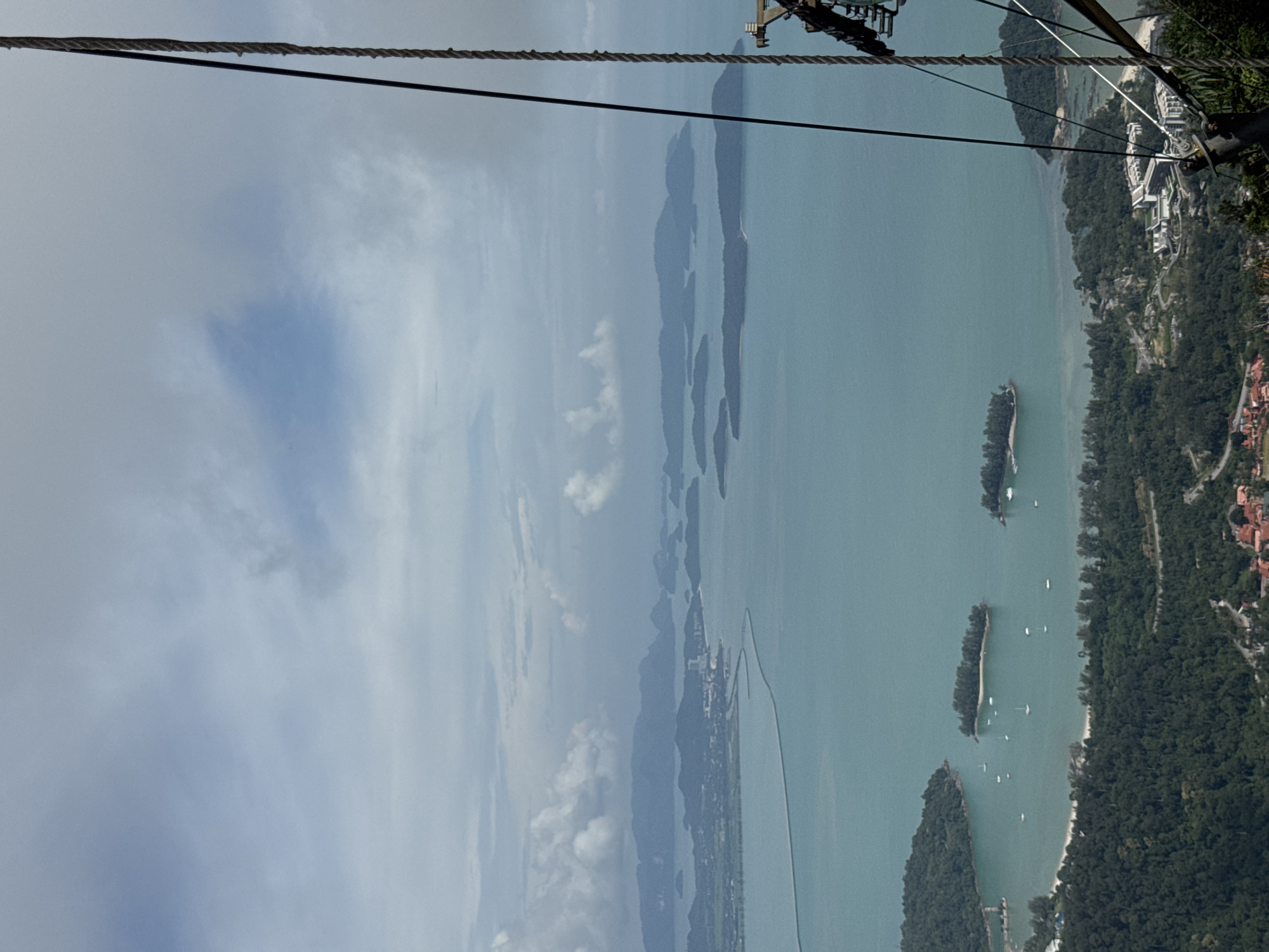 This is a high-angle, aerial view of a tropical island landscape. The foreground is dominated by the metallic structure and thick ropes of a cable car mechanism, suggesting the vantage point is from a cable car in motion or at a station. The scene looks out over a bay with turquoise waters dotted with numerous small islands covered in dense green vegetation. In the middle ground, a curved, man-made causeway or bridge stretches across the water, connecting a more developed part of the coastline to distant islands. This developed area appears to be a city or town with white buildings visible along the shore, nestled amongst the trees. The background features a hazy horizon with more mountainous islands fading into a cloudy sky. The overall impression is a panoramic view of a coastal area in Langkawi, Malaysia, likely taken from the popular Langkawi SkyCab. There are no people or animals clearly visible in the image.