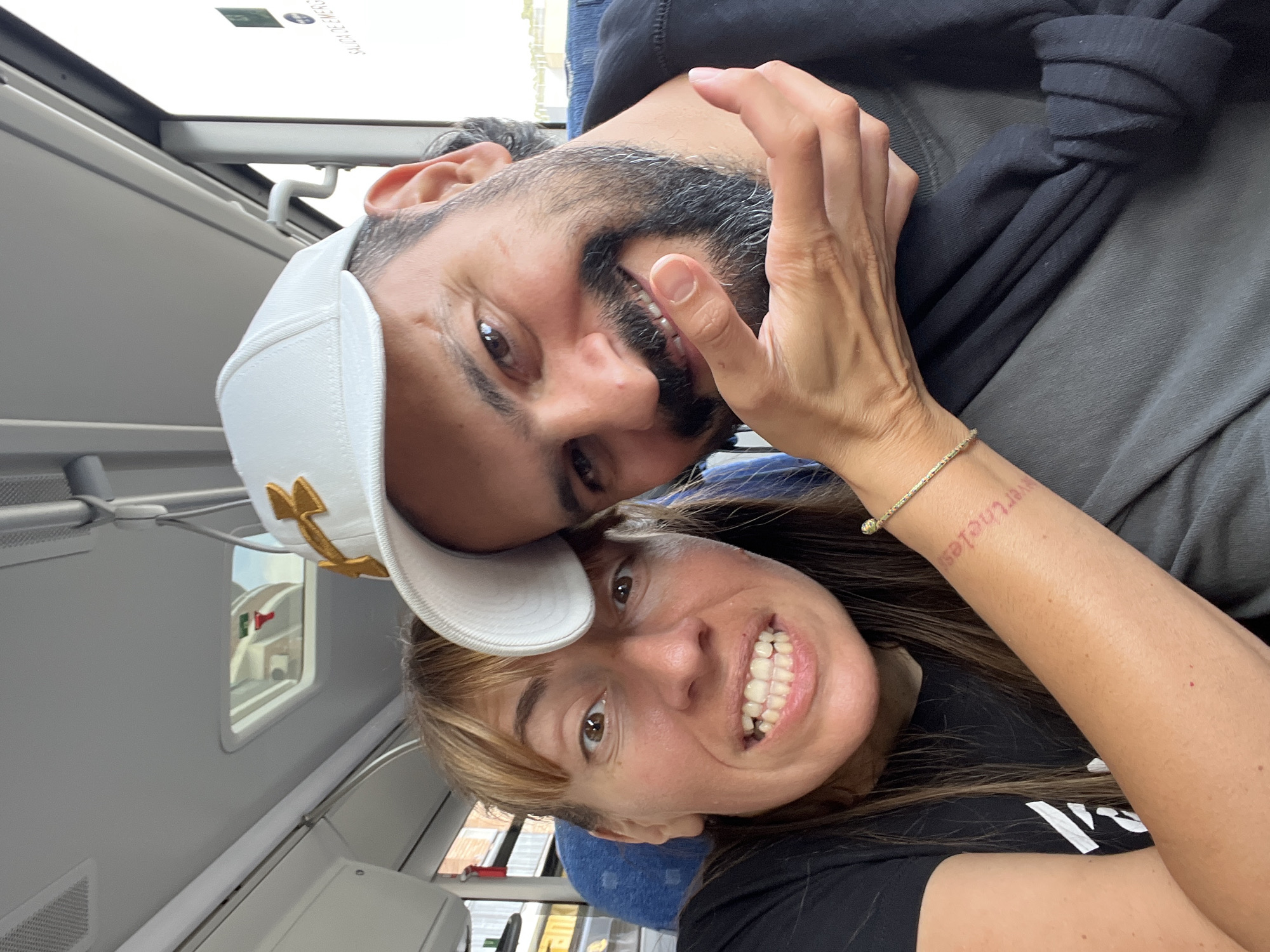 The image shows a close-up selfie of a man and a woman, appearing to be a couple, inside a vehicle. The man is wearing a white baseball cap with gold trim and has a beard. The woman has brown hair and is smiling broadly, showing her teeth. She has a tattoo on her forearm that reads "nevertheless". Both are looking at the camera. The background suggests they are seated in a bus or train, with windows visible. The lighting is bright, indicating it is daytime. The overall mood appears to be happy and relaxed, with the couple sharing a close moment.
