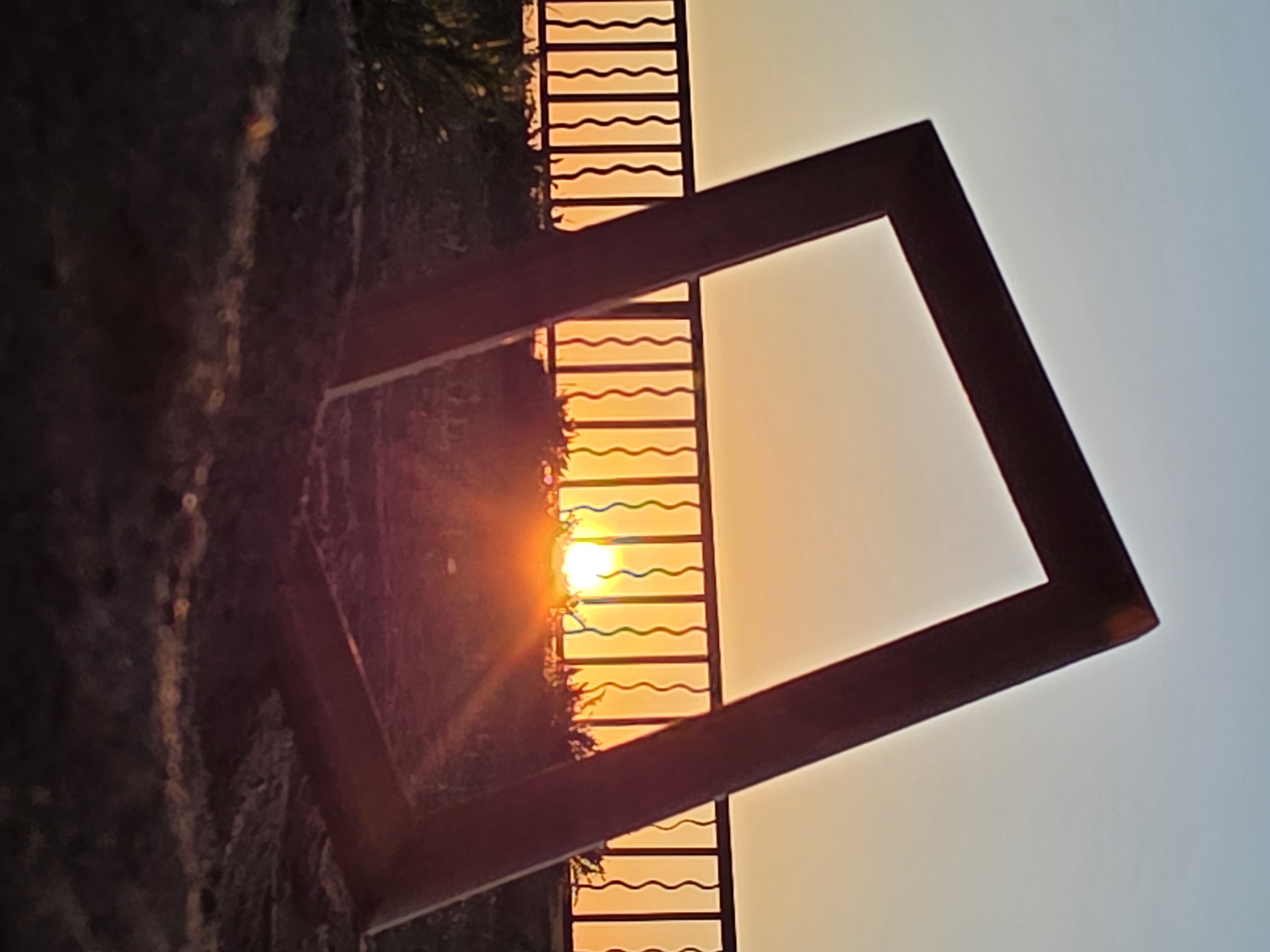 A photograph taken in Chaikovsky, Russia, features a skewed square frame positioned against a backdrop of a sunset viewed through a decorative metal barrier. The frame, appearing reddish-brown, is angled noticeably, creating a dynamic perspective. The sun, a bright focal point, is partially obscured by the barrier, radiating a warm glow. The metal barrier has a wavy pattern. The environment surrounding the frame and barrier includes foliage and what appears to be a wall. The sky transitions from orange near the horizon to a lighter hue above.
