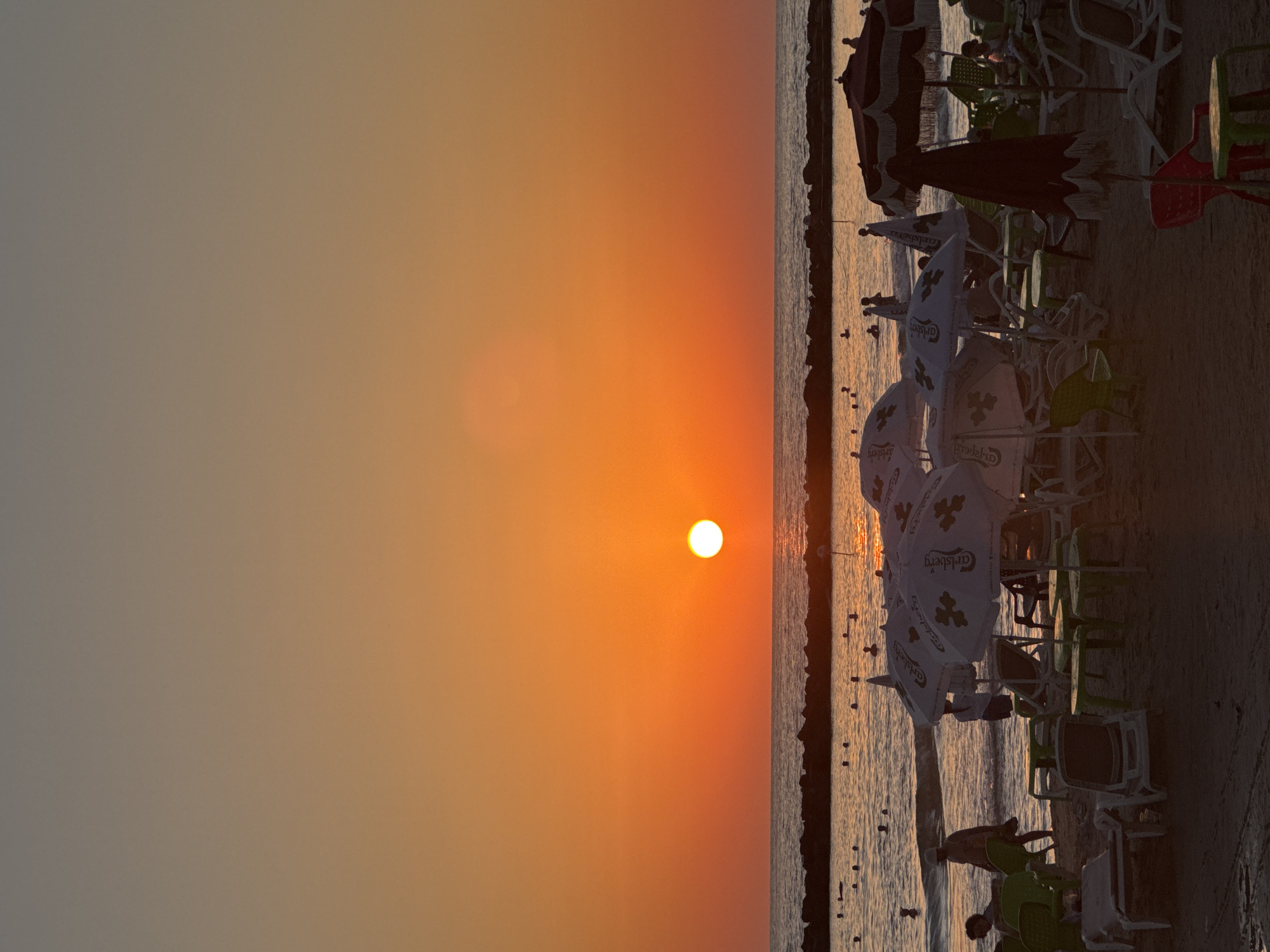 The image depicts a sunset scene on a beach in Netanya, Israel. The sun, a bright orange orb, is low on the horizon, casting a warm glow across the sky and water. The sea is calm, with a few scattered rocks forming a breakwater. Several people are visible in the water, swimming or wading. On the shore, beach chairs and umbrellas are set up, with some people sitting or walking on the sand. The overall atmosphere is peaceful and relaxing, characteristic of a late afternoon at the beach.