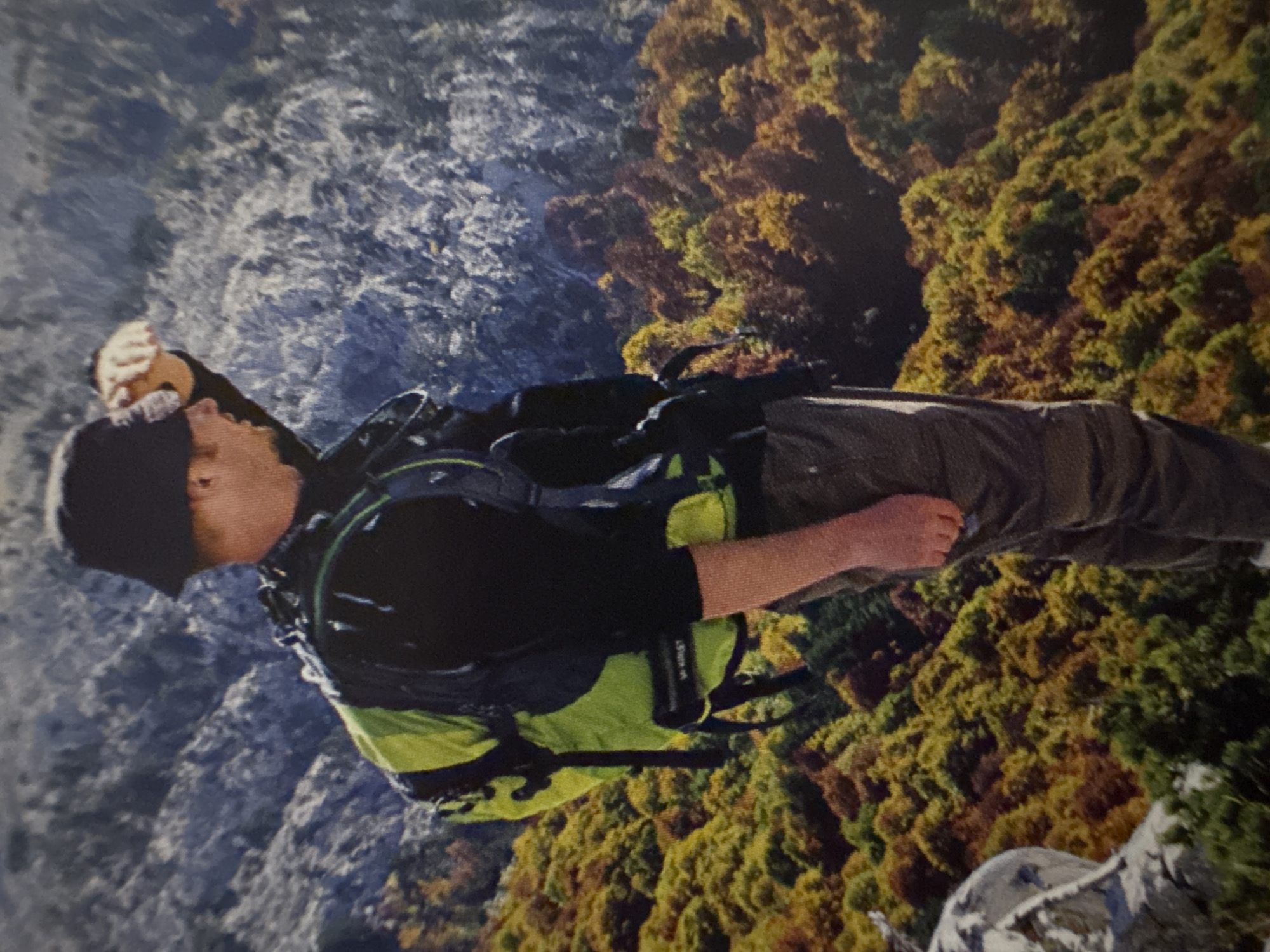 An image shows a man wearing a black bucket hat and a black shirt, standing in front of a mountain range. He has a lime green backpack on and is looking into the distance, with his hand raised to his forehead. The mountain range appears to be covered in trees that are changing color, suggesting autumn. The image appears to be taken from a high vantage point, looking down at the man and the landscape. The man's lower body is visible, wearing grey trousers. The background shows rocky, grey mountains with a forest of trees displaying shades of green, yellow, orange, and red.