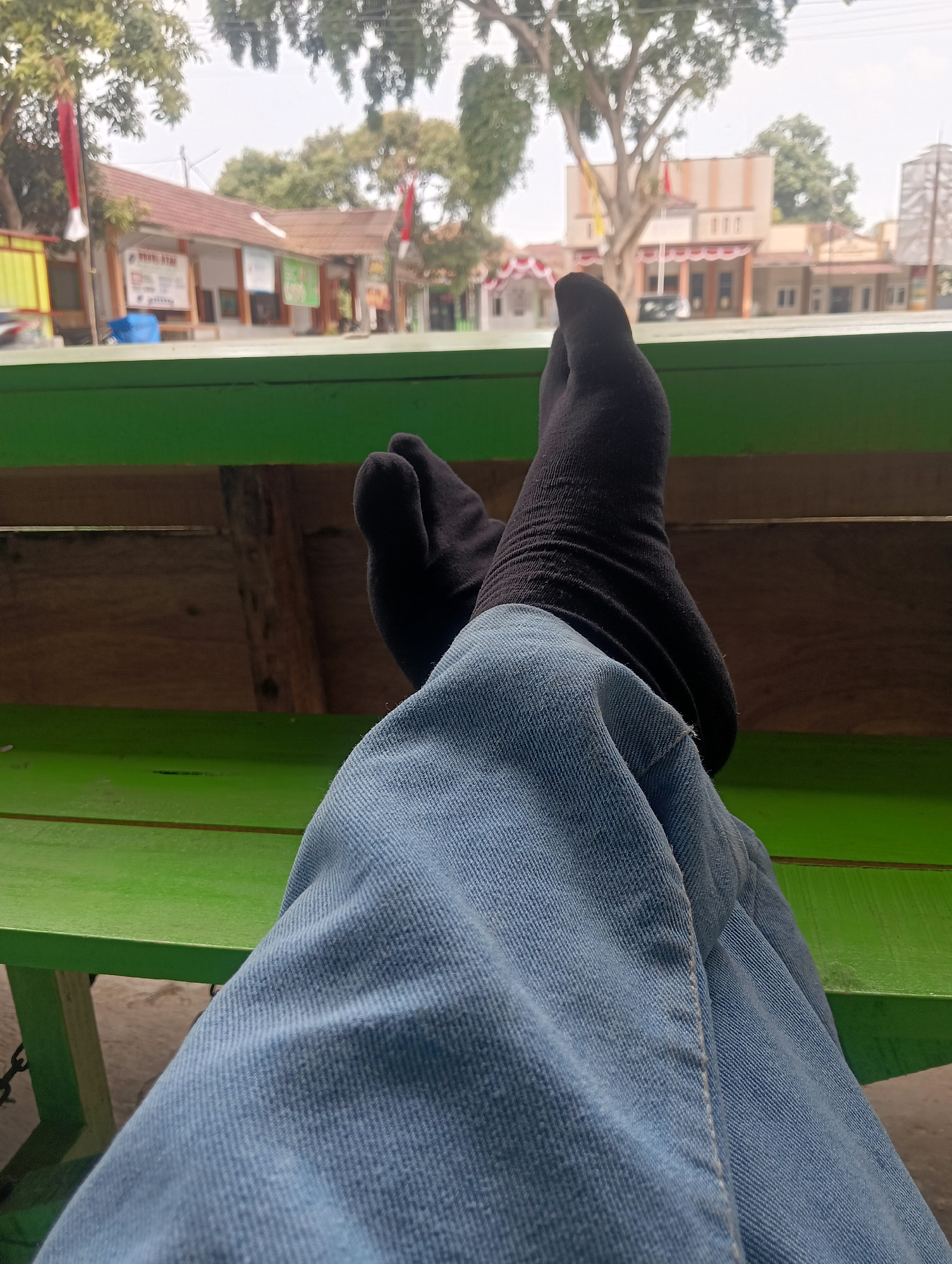 The image shows a close-up perspective of a person's legs, clad in blue jeans and black tabi socks, resting on a green bench. The background, slightly out of focus, depicts a street scene in Manggari, Indonesia, with buildings, trees, and flags visible. The architecture of the buildings suggests a tropical or sub-tropical climate. The overall impression is one of casual relaxation in an outdoor setting.