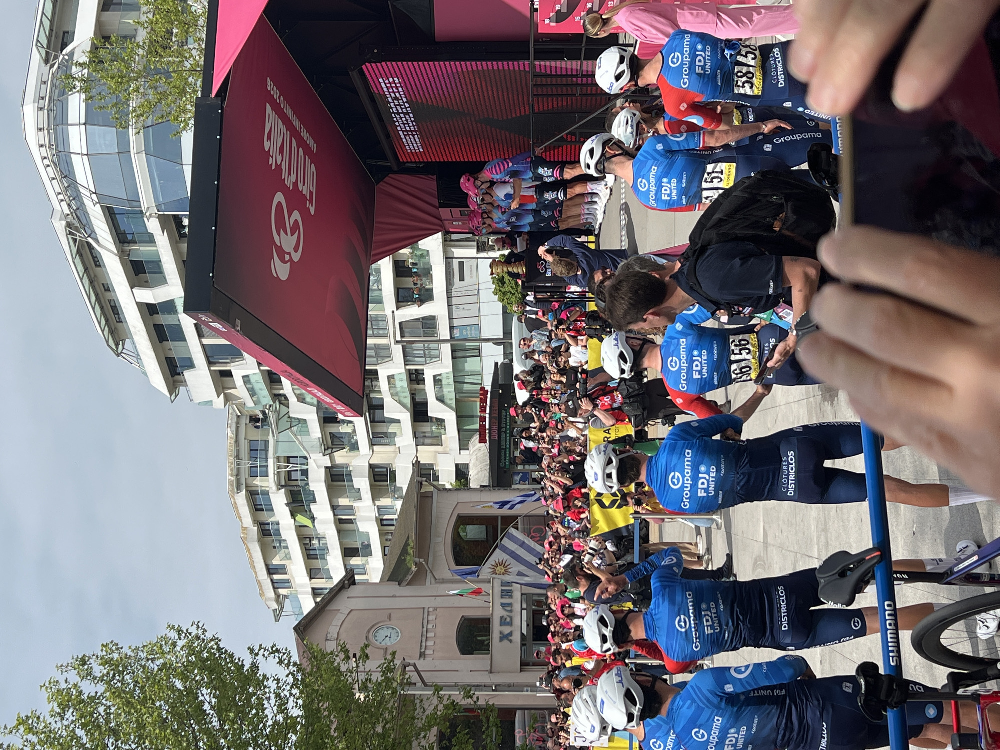 The image captures a scene at the Giro d'Italia cycling event in Burgas, Bulgaria. Cyclists, wearing blue Groupama-FDJ United jerseys with helmets and numbered bibs, are standing along a barrier. Spectators crowd behind the barrier, some holding flags of Bulgaria and Uruguay. In the background, there's a stage with a Giro d'Italia banner. A modern white building with a glass facade towers over an older pink building with a clock tower and the word "ΧΕΛΗ" written on it. The photographer is holding a smartphone, capturing the event. Bike equipment like handlebars and a Shimano pedal can be seen.