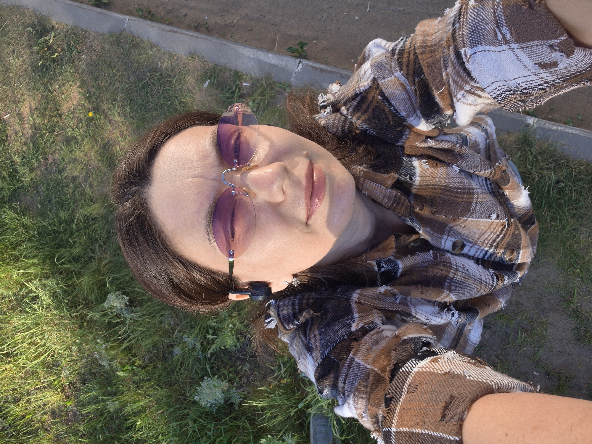 A woman lies on her back in a grassy area beside a sidewalk. She wears rimless tinted sunglasses and a brown and white plaid shirt. The grass is green and interspersed with various plants. A concrete sidewalk edge is visible in the background.
